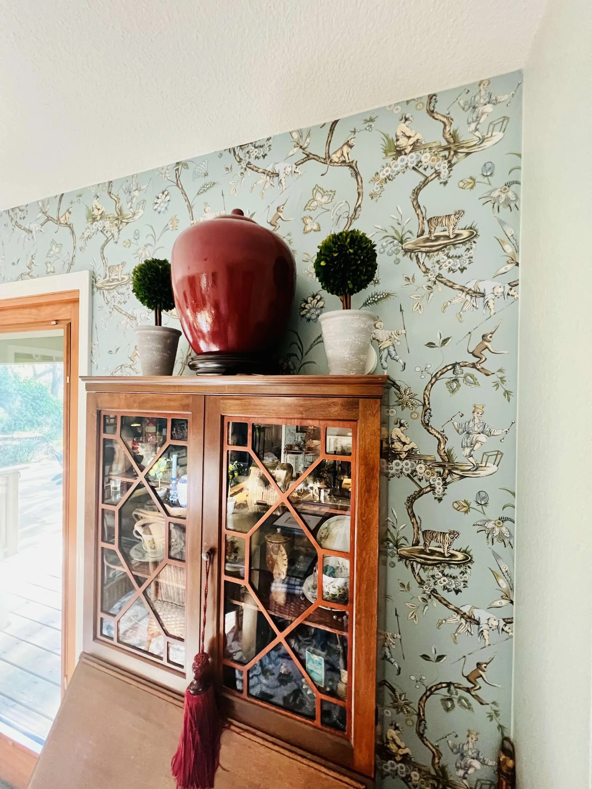 A wooden display cabinet with glass panes and a geometric pattern, topped with a large red ceramic vase and two potted topiary plants, against a wallpapered wall with a sky-blue background and illustrations of people, animals, and plants.
