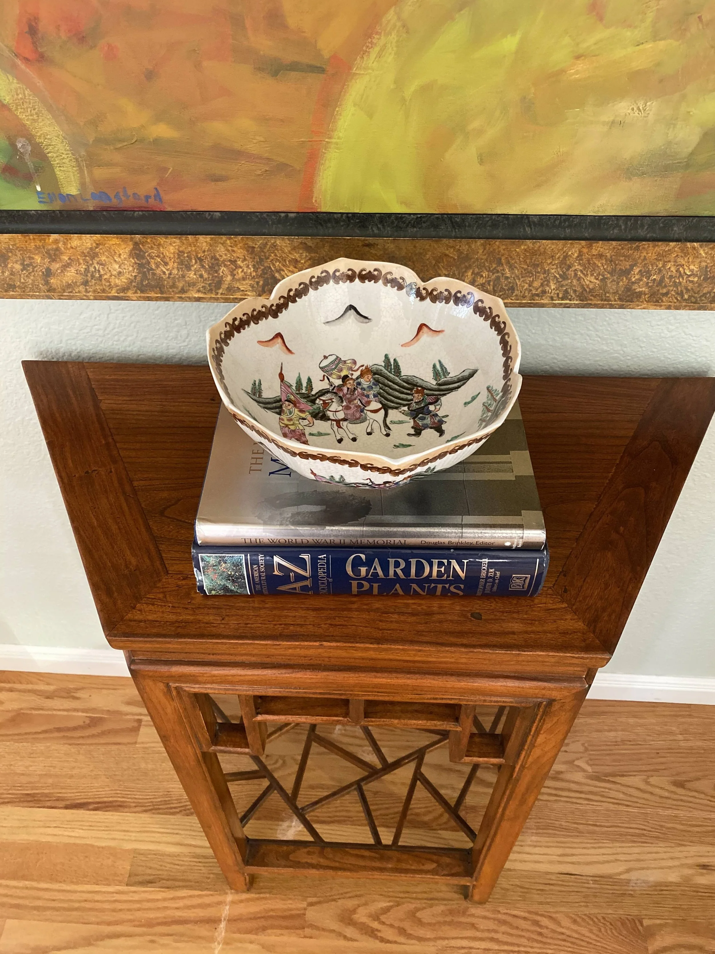 A wooden side table with an open-bottom lattice shelf beneath a marble top, on which two hardcover books are stacked. The top book is titled 'The World War II Memorial' and the bottom book is titled 'Garden Plants'. A decorative ceramic bowl with a painted scene of people riding horses is placed on top of the books. In the background, a colorful abstract painting hangs on the wall.