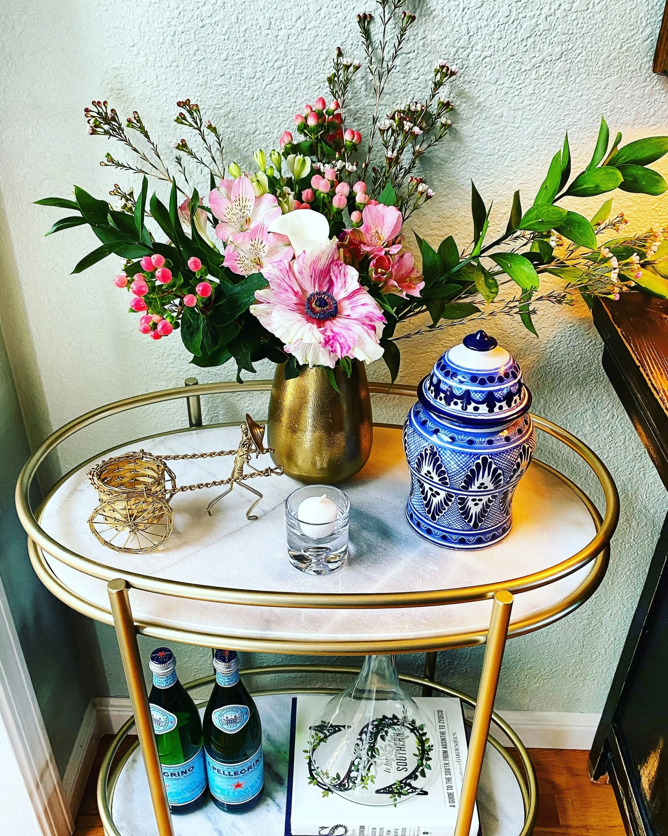 A gold-colored metal stand with a white marble top holding a floral arrangement with pink, white, and green flowers and greenery, a blue and white patterned decorative jar, two bottles of San Pellegrino sparkling water, a glass of water, a tealight candle in a glass holder, a golden wire decorative sphere, and a tray underneath with what appears to be a booklet with a black design.