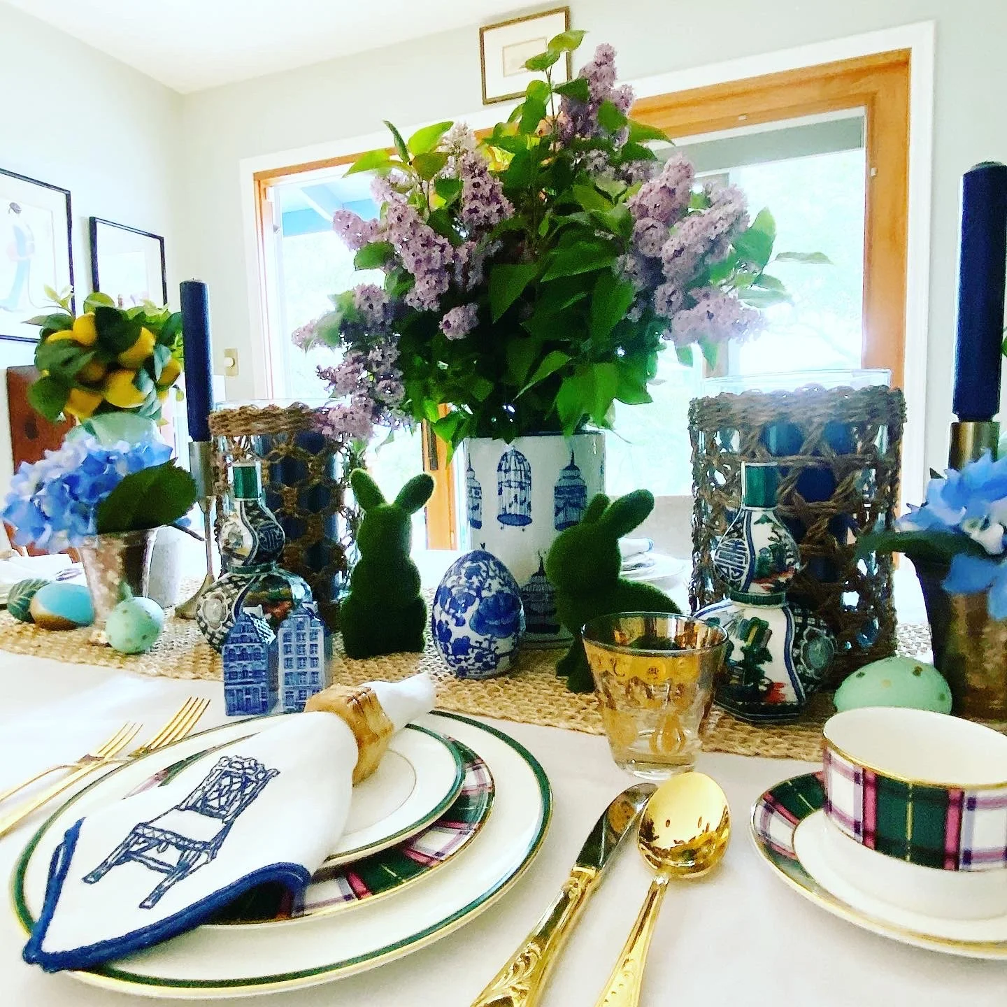 Table setting with decorative Easter eggs, gold utensils, and themed dinnerware. The background features floral arrangements, bunny figurines, and decorative vases, creating a festive spring or Easter decoration.