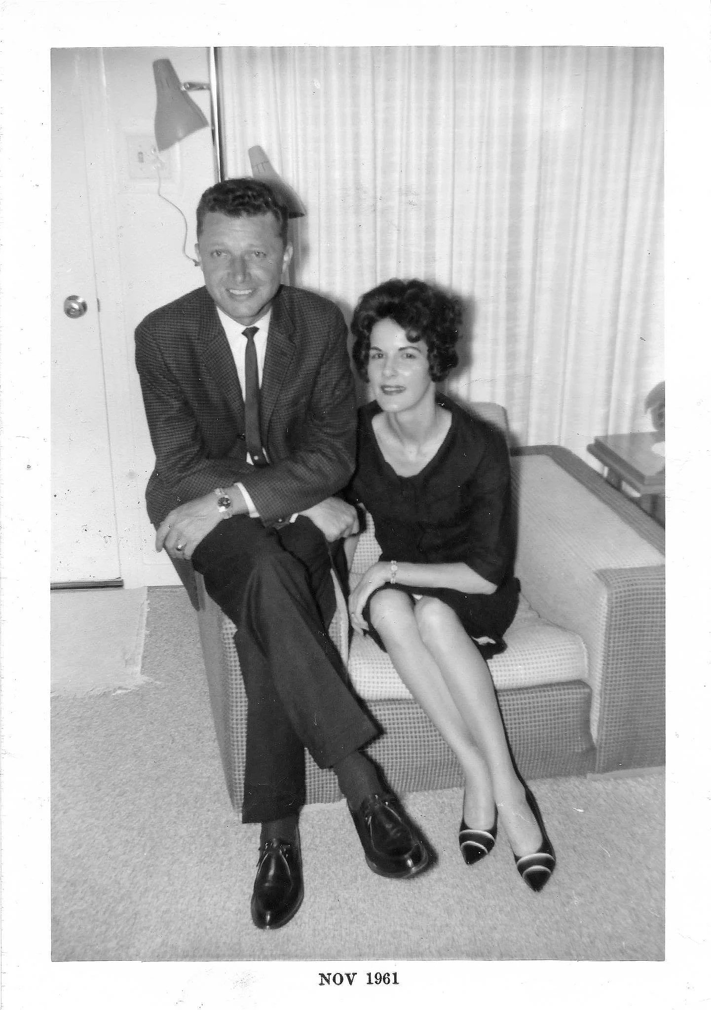 A man in a suit with a woman in a black dress sitting on a bench in a room with curtains. The man has short hair and is smiling; the woman has curly hair and is looking at the camera. The photo is black and white, dated November 1961.