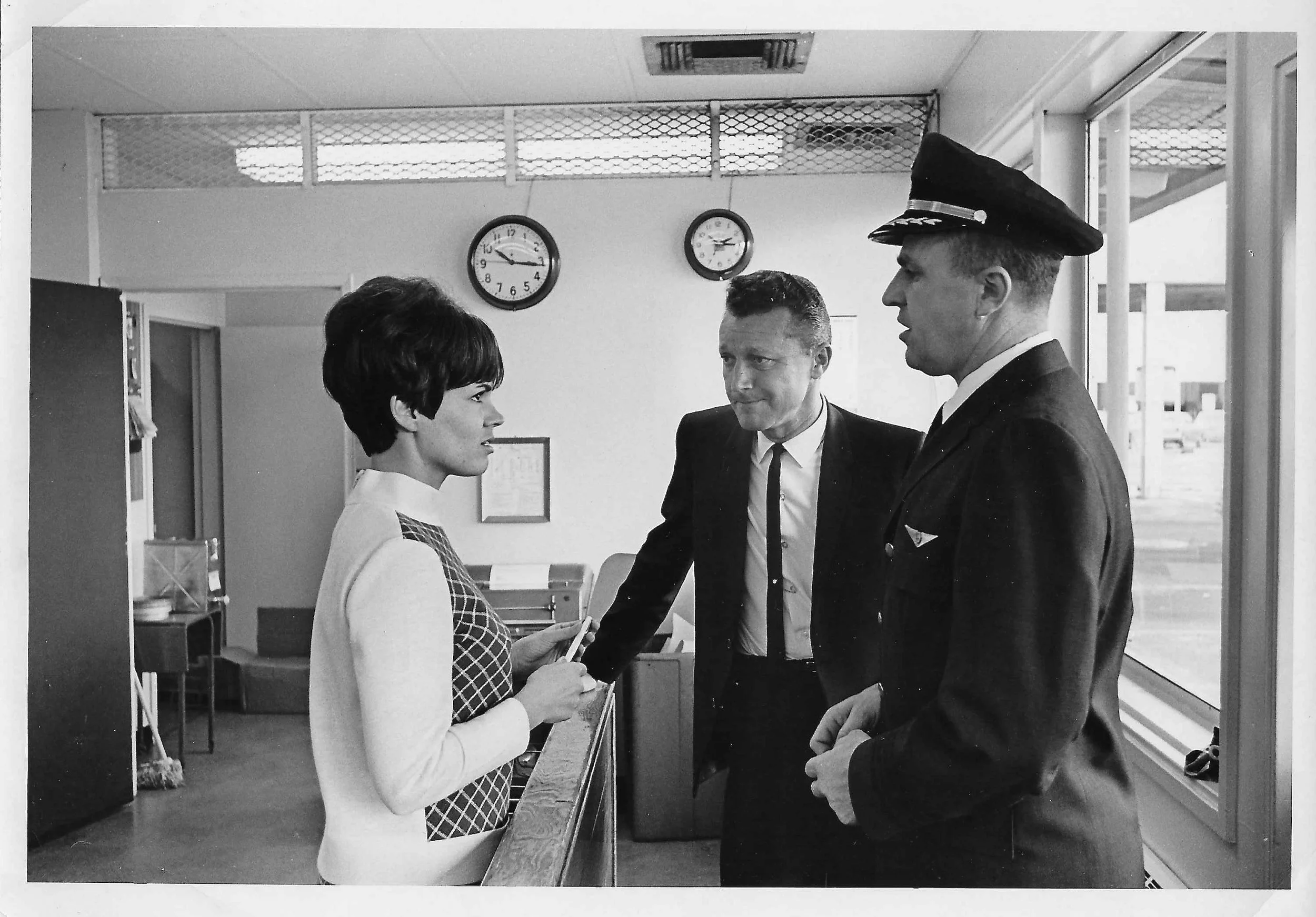 A black and white photo showing a woman and two men inside an airport or train station. The woman is holding a boarding pass and is engaging in conversation with one man, who is wearing a police or military uniform. The other man is dressed in a suit and tie, standing between them. There are clocks on the wall and a doorway in the background.