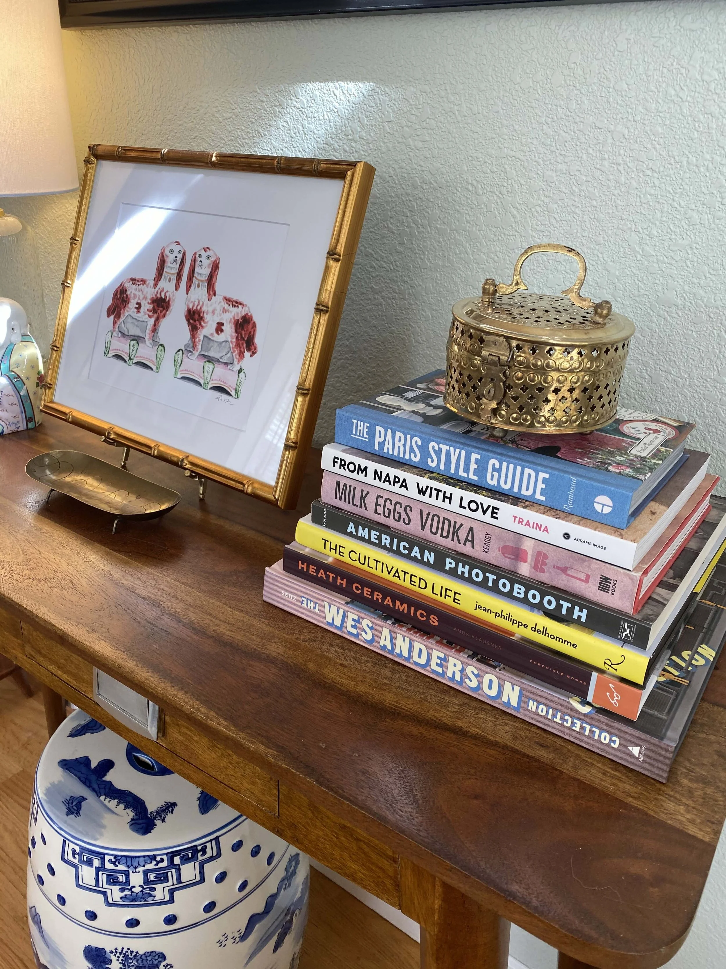 A wooden table with a stack of interior design and travel books, a decorative gold container, a framed watercolor artwork of two dogs, a modern lamp, and a porcelain vase with blue patterns, in a well-lit room.
