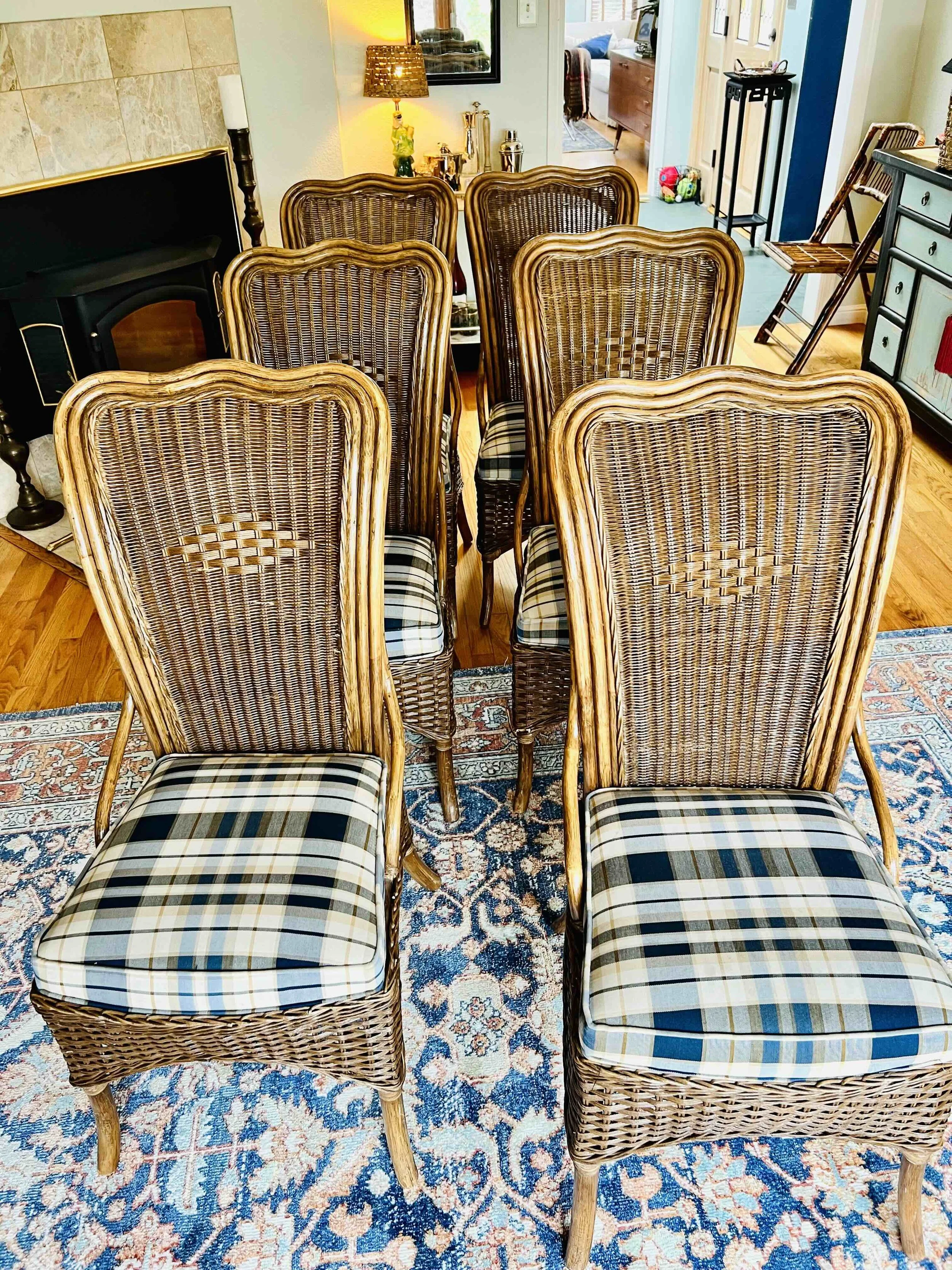 Six chairs with woven rattan backs and plaid cushioned seats arranged in a room with a patterned rug. In the background, there are various decorative items including a lamp, a black fireplace, and a hallway leading to other rooms.
