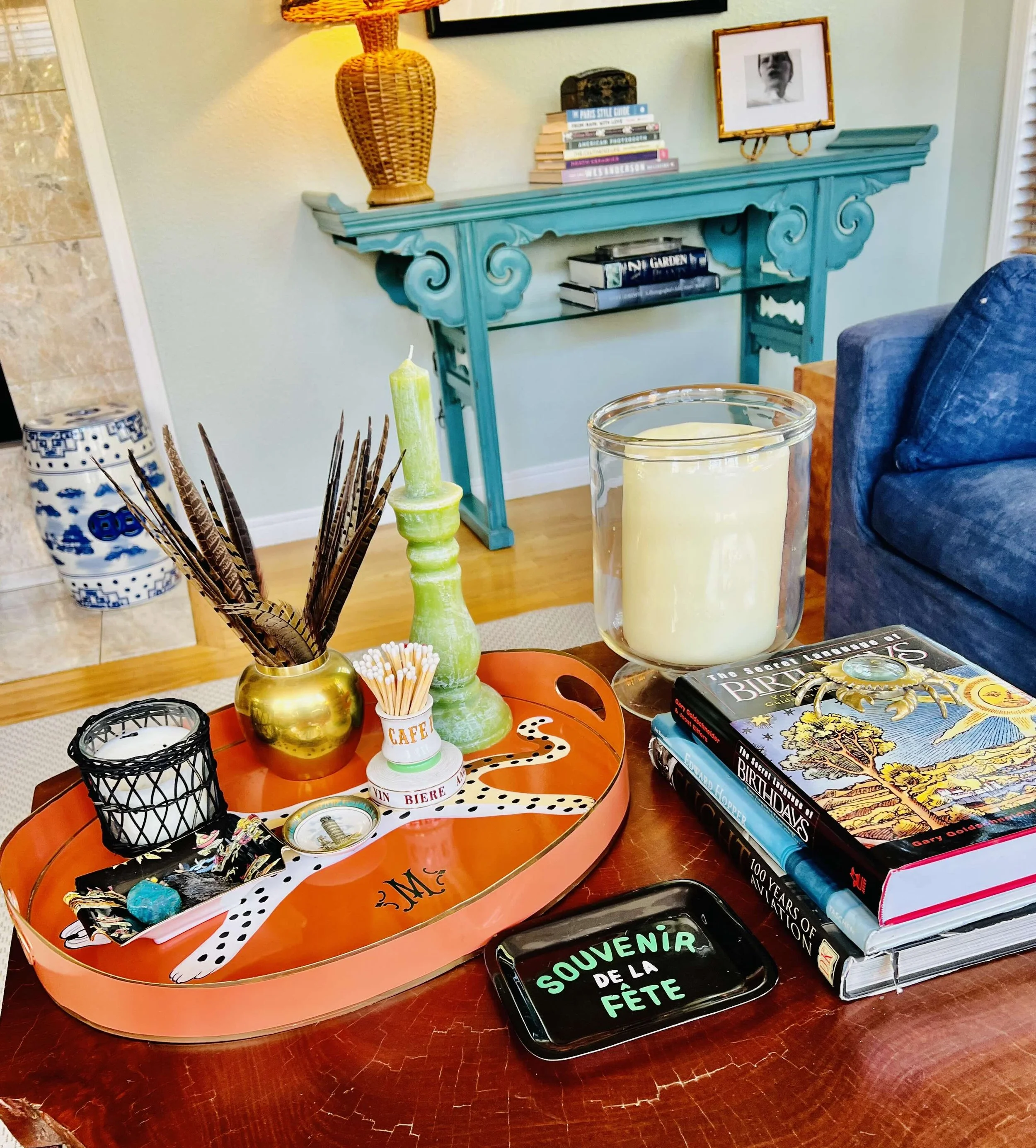 Decorative living room with teal-colored carved fireplace mantel, decorated with a wicker lamp, stacked books, and framed black-and-white photos. The foreground features a coffee table with candles, a tray with a painted leopard print, and books about birthdays and celebrations.