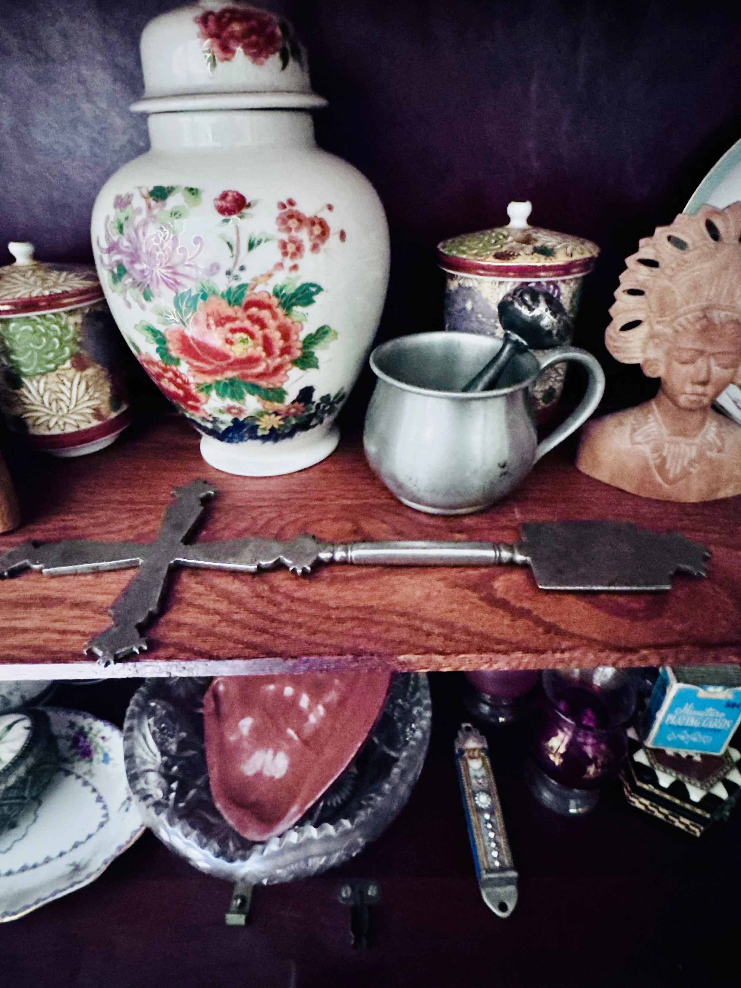 A collection of vintage and decorative items displayed on a wooden surface, including floral ceramic jars, a metal mug with a spoon, a carved wooden figurine, and various assorted decorative objects.