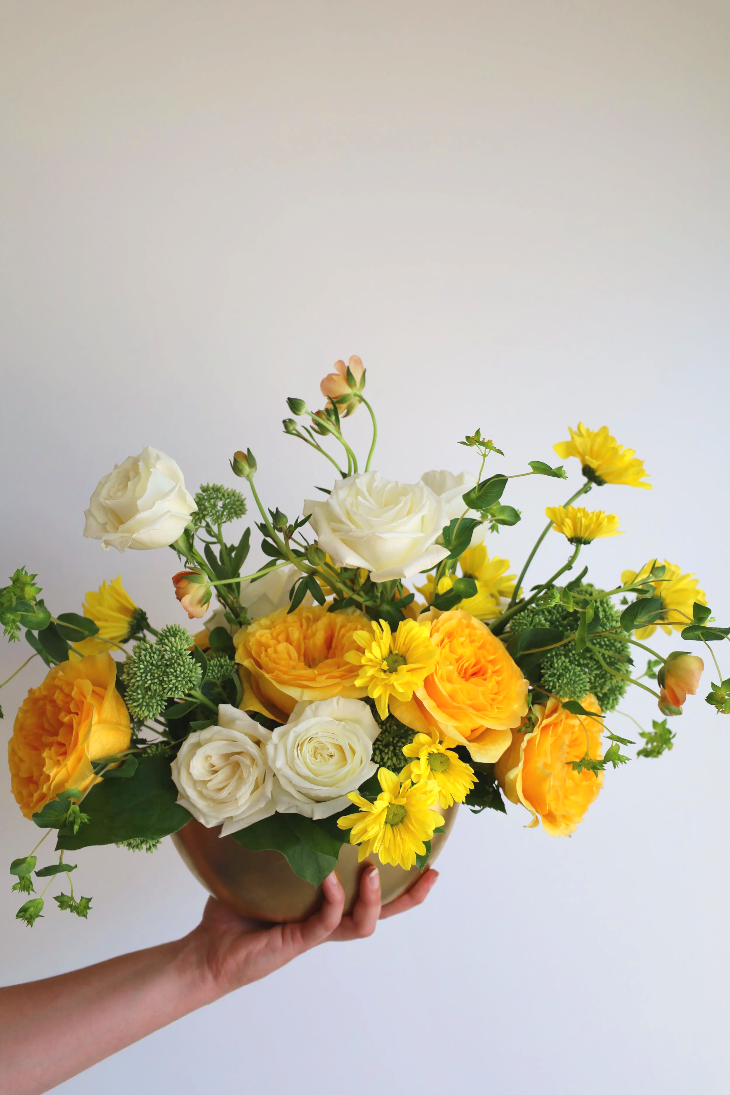An arrangement of white and yellow flowers in a gold vase against a plain light background.