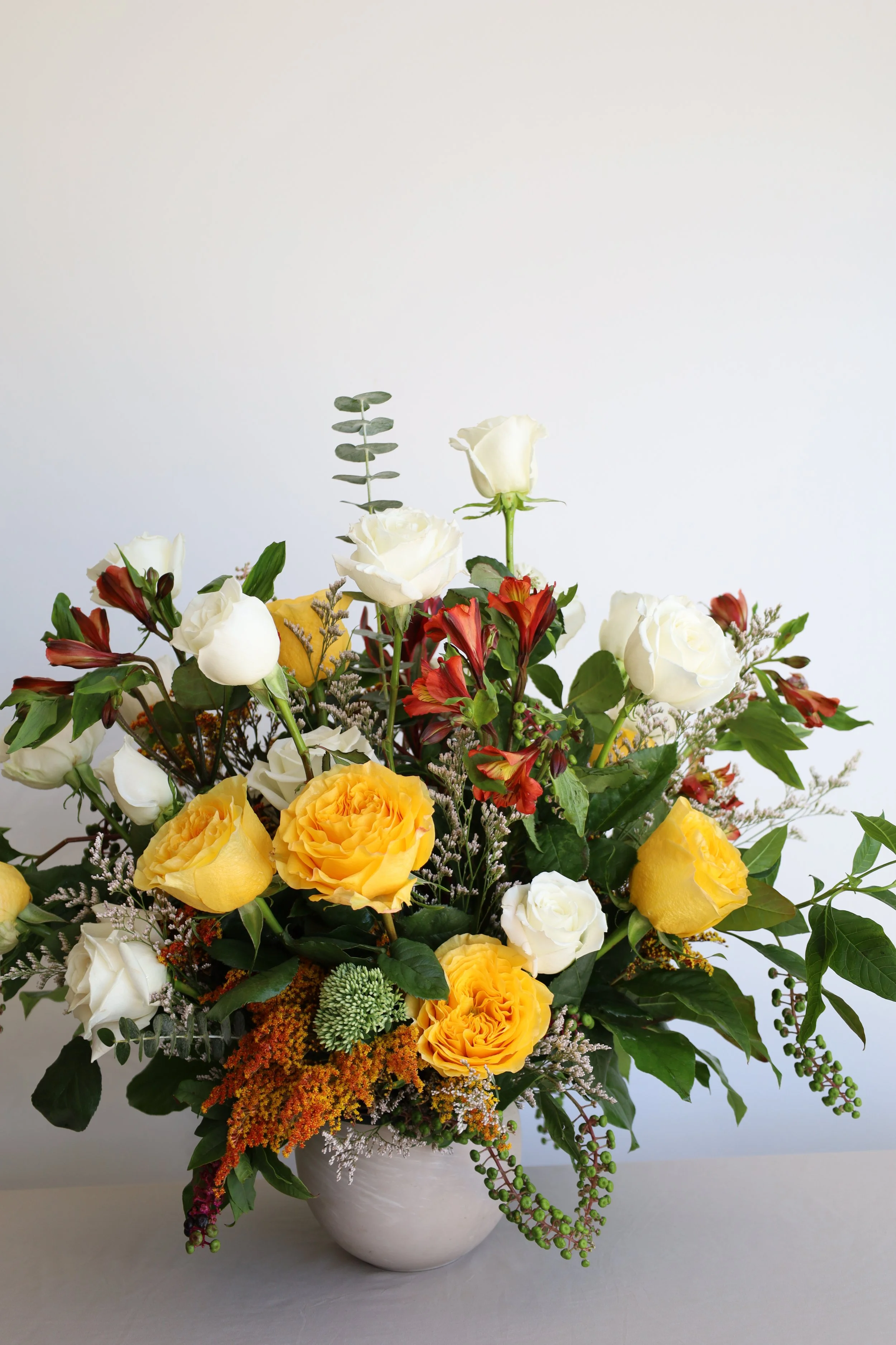 A large floral arrangement in a clay vase, featuring yellow roses, white roses, orange flowers, and greenery, set against a white background.