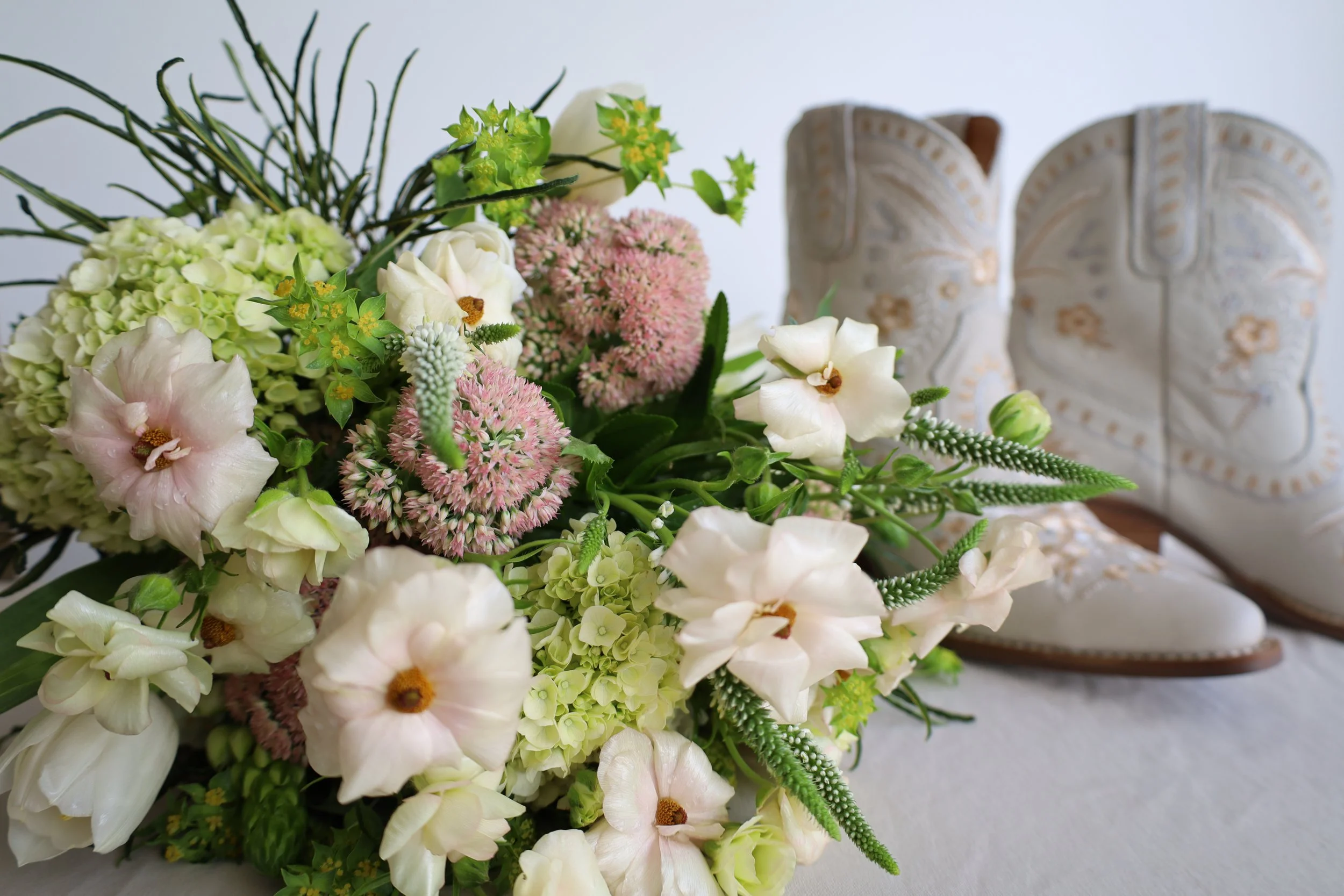 A colorful bouquet of various flowers including white roses, pink hydrangeas, and greenery, with a pair of embroidered white cowboy boots placed in the background.