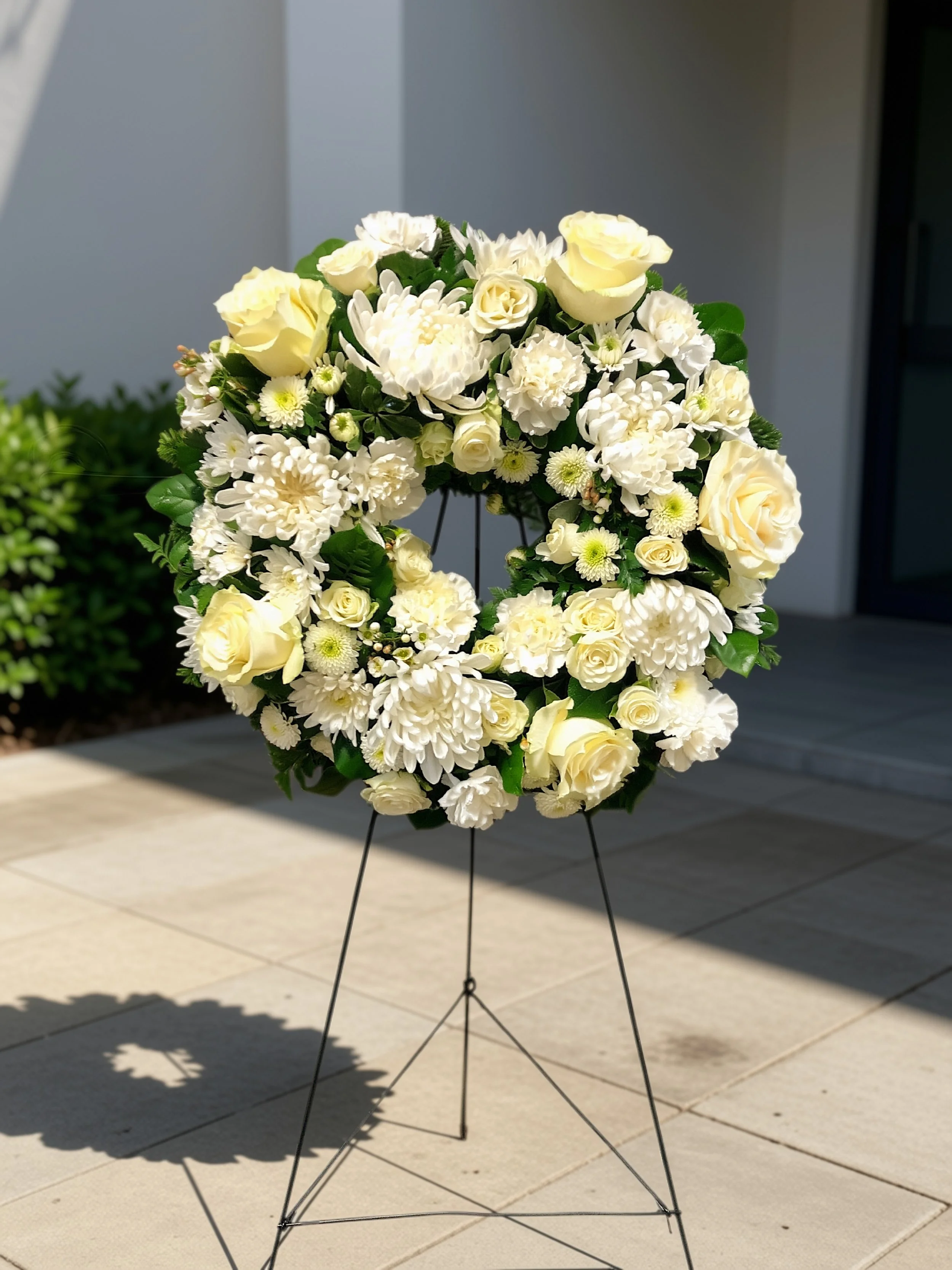 White floral wreath on a stand with a black metal base, set outside on a tiled surface.