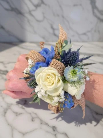 A wrist corsage with white roses, blue flowers, greenery, and burlap accents, against a marble background.