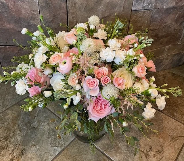 A bridal bouquet of pink and white roses, ranunculus, other flowers and various greenery in a vase on a tiled floor.