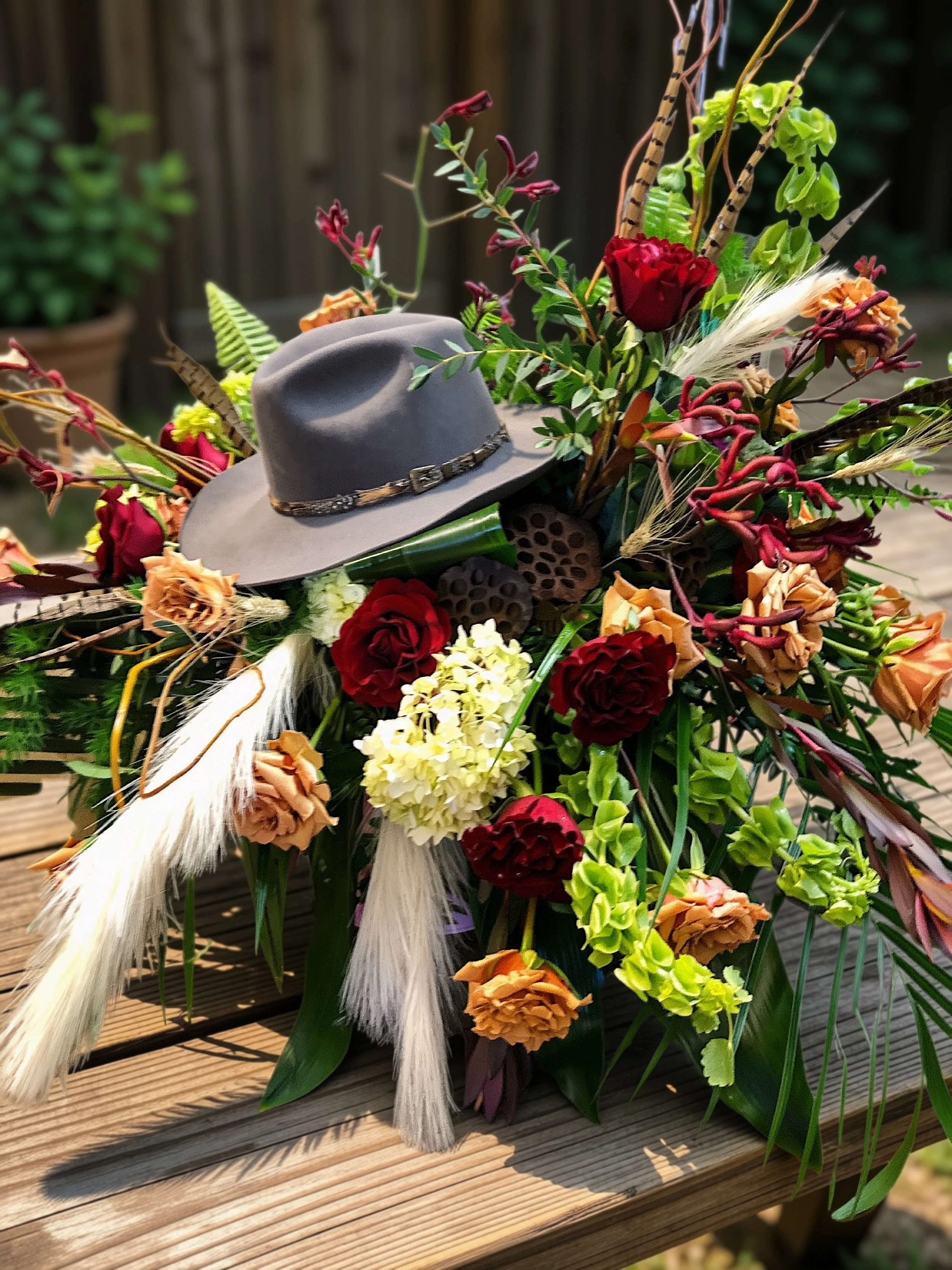 A floral arrangement with various flowers, dried grasses, and a small gray hat with a band, placed on a wooden surface.