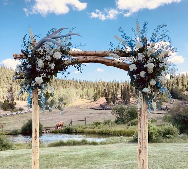 Wooden wedding arch decorated with white and blue flowers and antlers, set outdoors near a pond with trees in the background.
