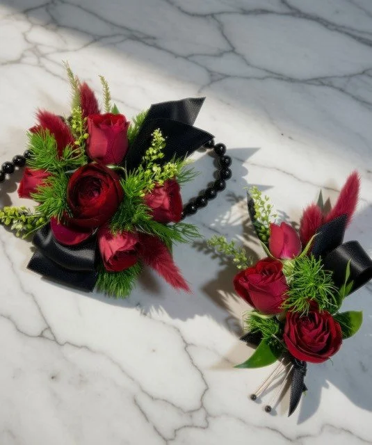 A wrist corsage and a boutonniere, featuring red roses, greenery, and black ribbons, resting on a marble surface.