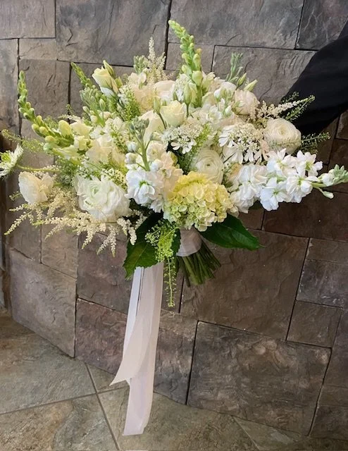 A large bridal bouquet of white and cream flowers with green foliage, wrapped with white trailing ribbons against a gray stone wall.