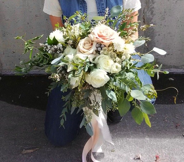 Person holding a large bridal bouquet of white and cream roses, other flowers and greenery, with a white and cream trailing ribbon.