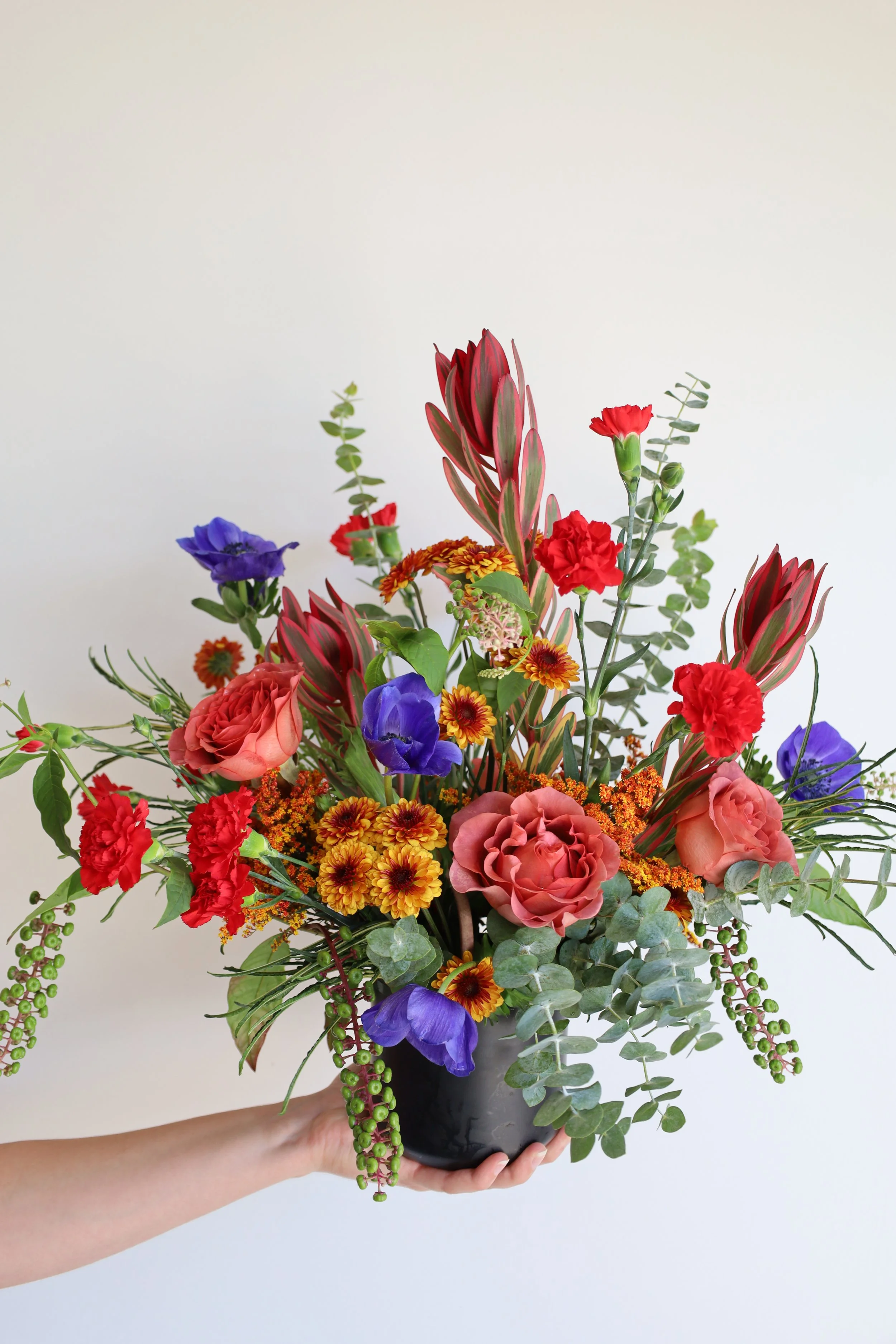A hand holding a black vase filled with a colorful arrangement of flowers and greenery against a plain background.
