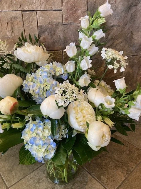 An arrangement of white and light blue flowers arranged in a glass vase, set against a stone wall.