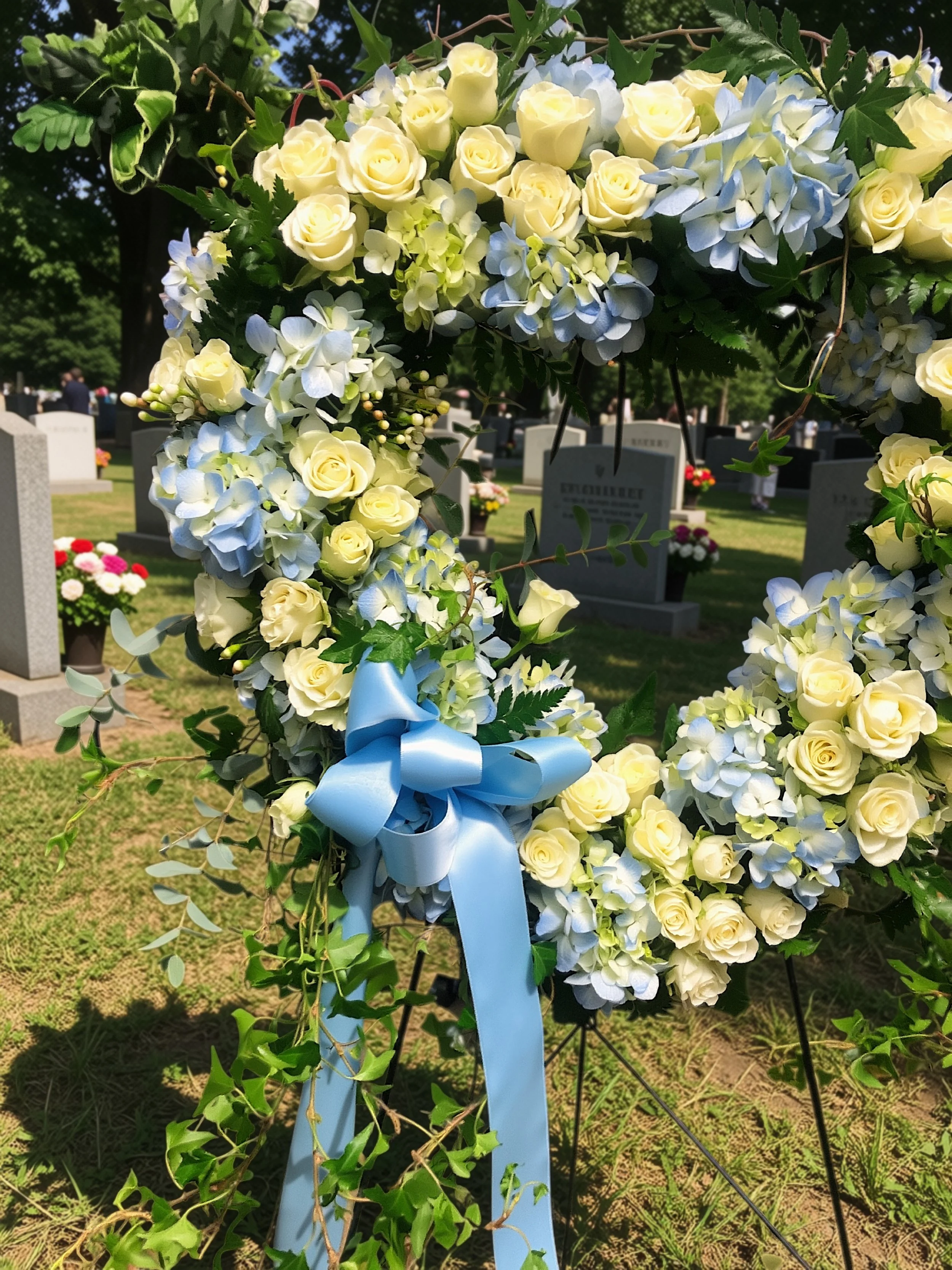 A floral open heart with white roses, light blue hydrangeas, and greenery, adorned with a blue ribbon.