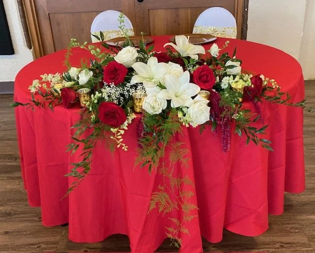 A round table covered with a red tablecloth, decorated with a floral arrangement consisting of white lilies, red roses, white roses, and greenery. Two chairs are positioned behind the table, with white covers and gold accents on the chairs.