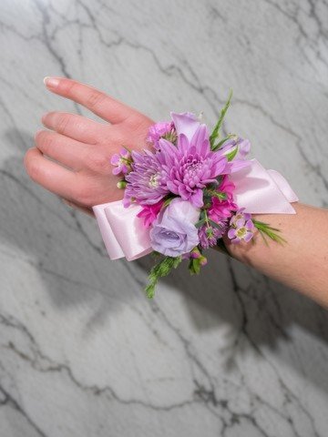 A person's wrist with a corsage of pink and lavender flowers and a pink ribbon against a light gray marble background.