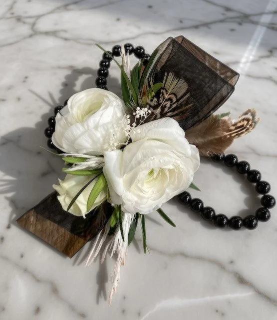 A wrist corsage with white ranunculus, greenery, feathers, and a sheer black ribbon, set on a marble surface.