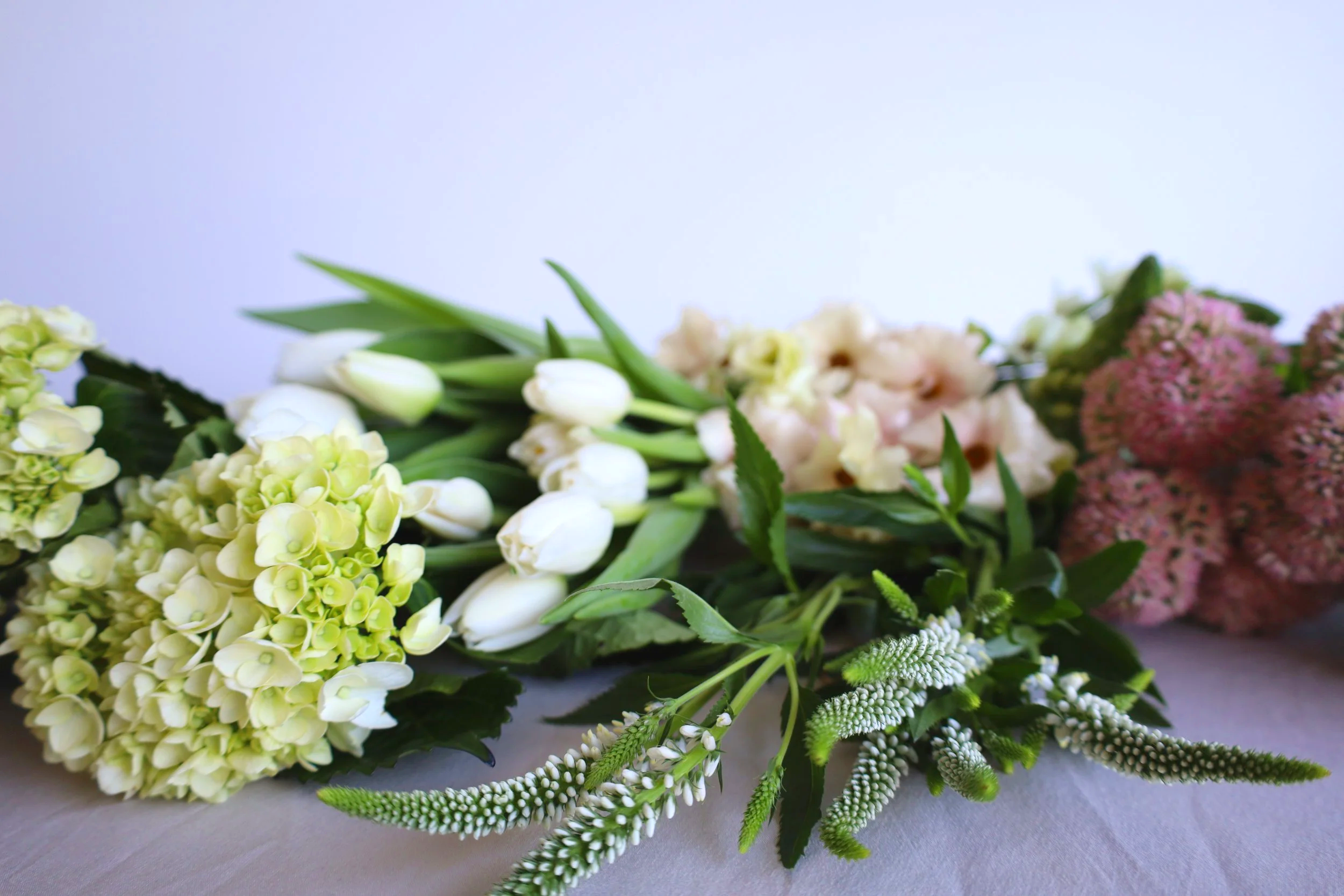 An assortment of fresh flowers including white tulips, green hydrangeas, pink and white flowers, and various green leaves arranged on a surface.