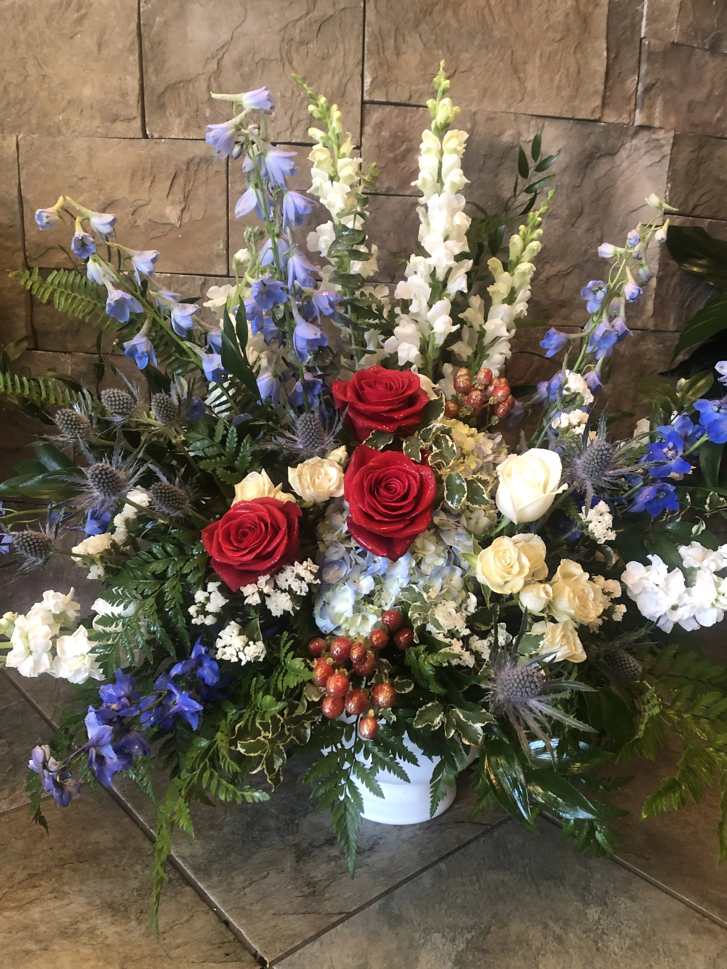 A sympathy arrangement in a white container with red, white, and blue flowers with greenery, placed on a tiled floor against a stone wall background.