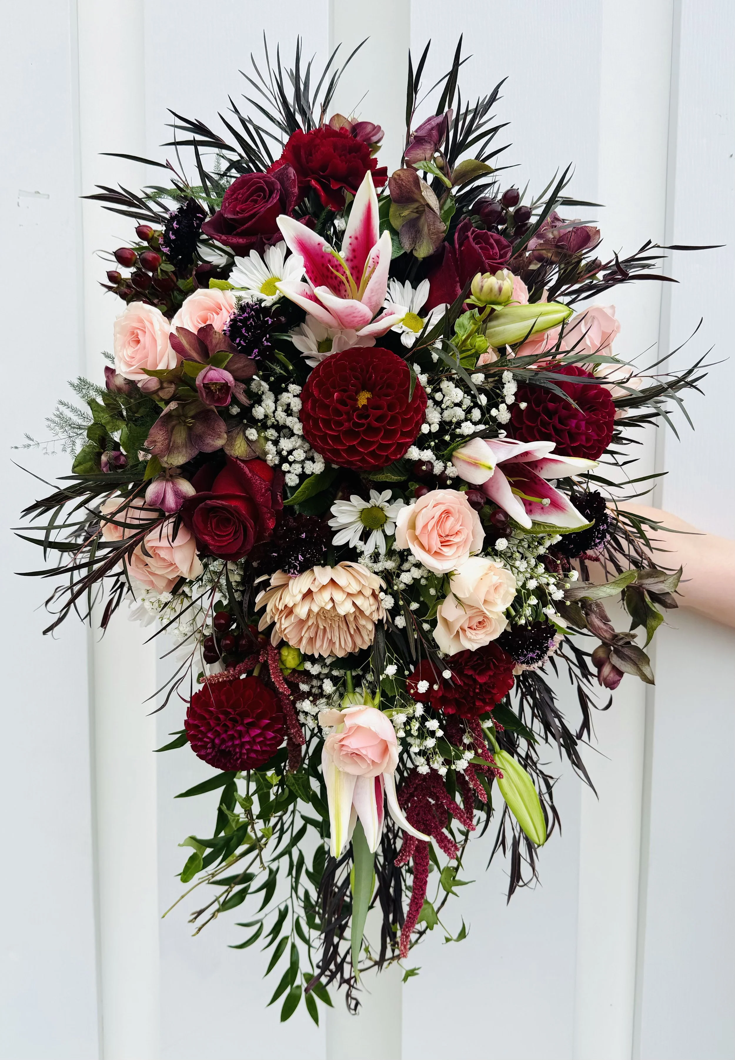 A floral bouquet with red roses, pink lilies, white daisies, and dark purple flowers, accented with greenery and dark leaves against a white background.