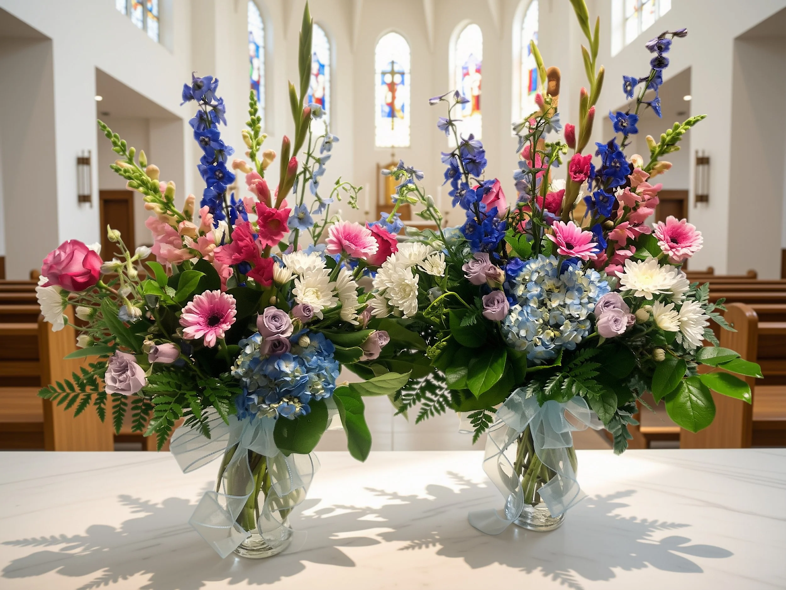 Two glass vases filled with colorful fresh flowers, including pink, white, and blue blooms, placed on a white table inside a church with stained glass windows.