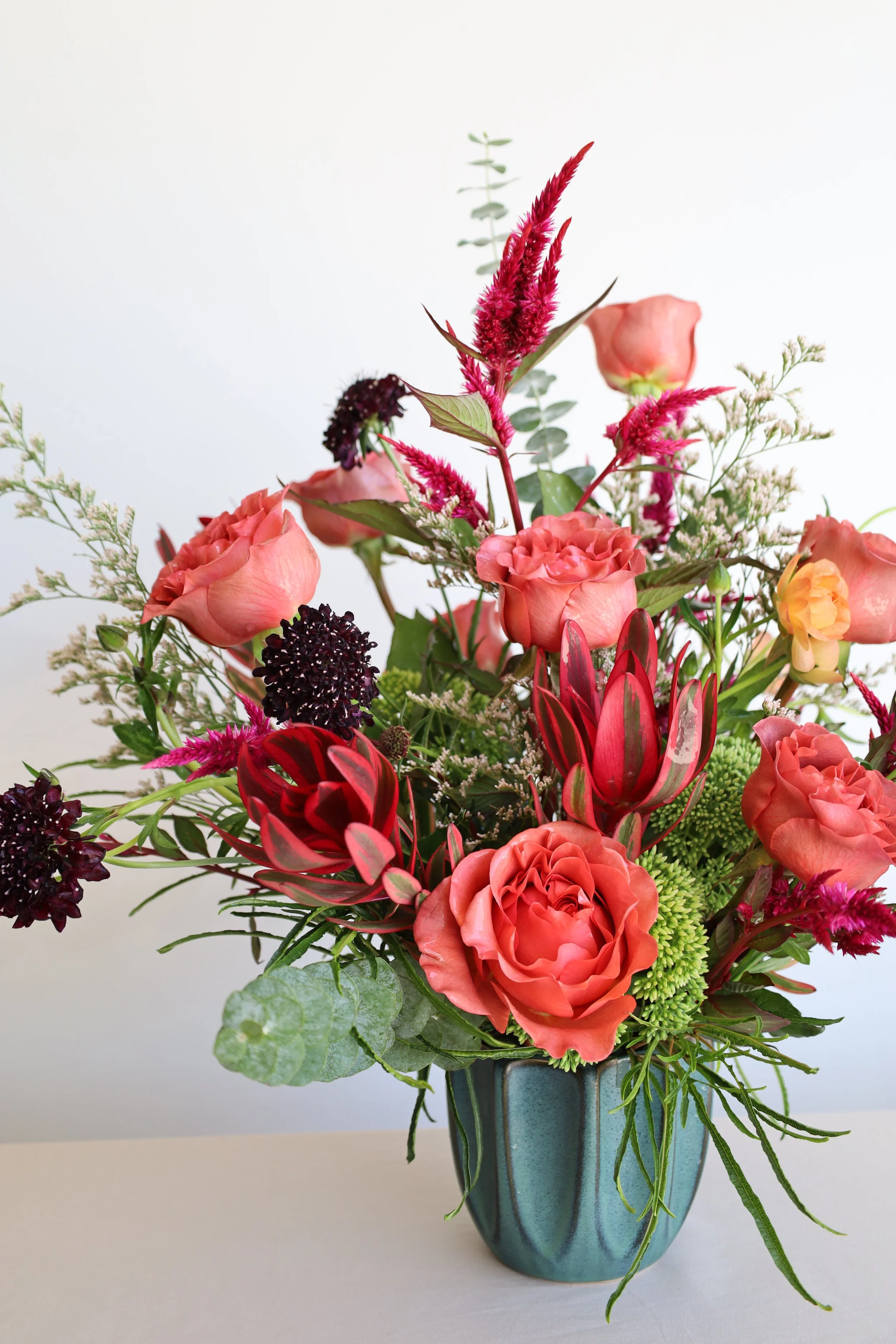 A colorful flower arrangement in a blue-green ceramic vase on a white surface, against a white background.