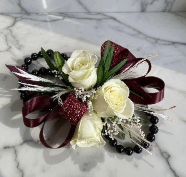 A wrist corsage with white roses, burgundy ribbon, greenery, and dried accents, placed on a marble surface.