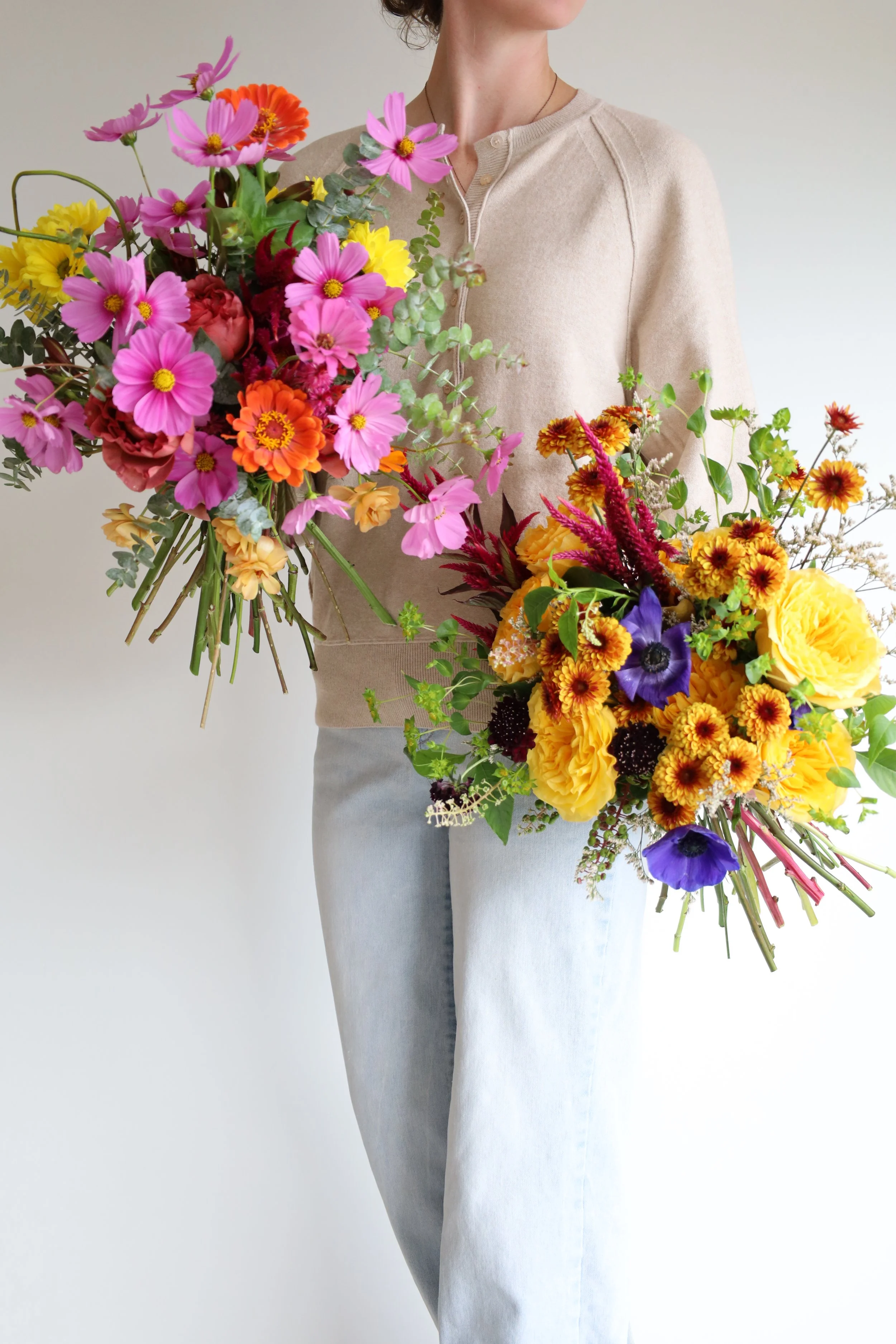 Person holding colorful bouquets of flowers, with beige top and light blue jeans.