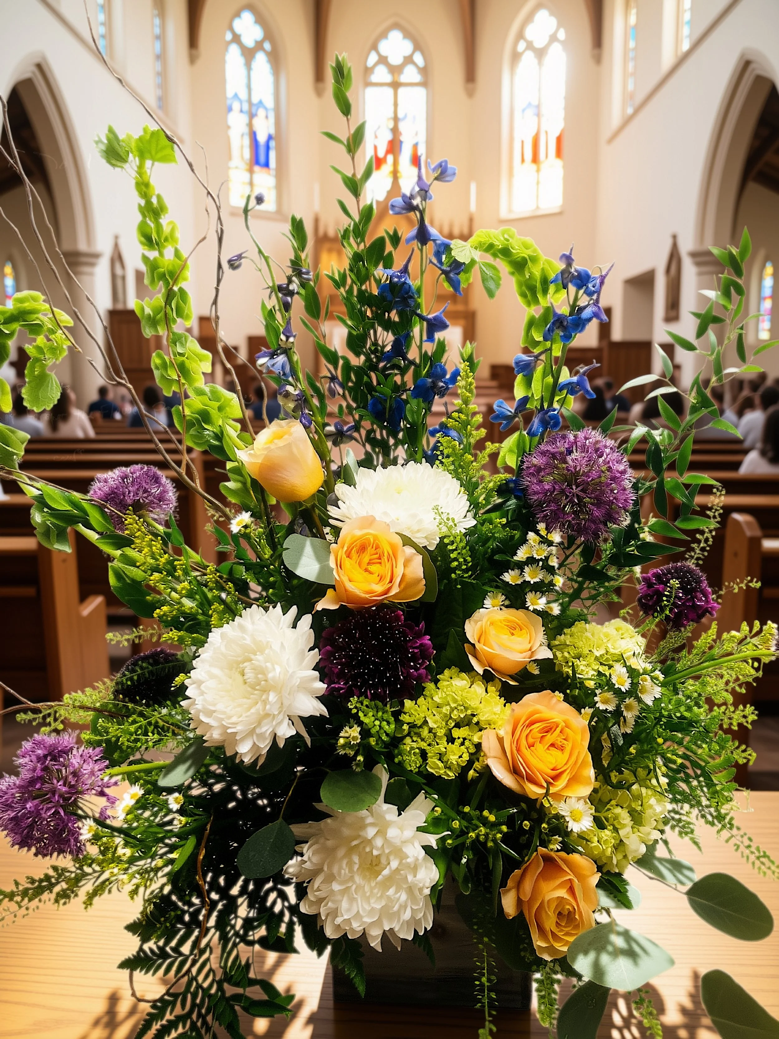 A colorful flower arrangement with yellow roses, purple and blue flowers, white mums, and greenery in a black container inside a church with tall stained glass windows.