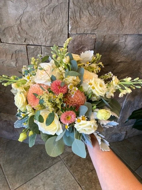 Hand holding a bridal bouquet of peach, cream, and yellow flowers with eucalyptus accents against a stone wall background.