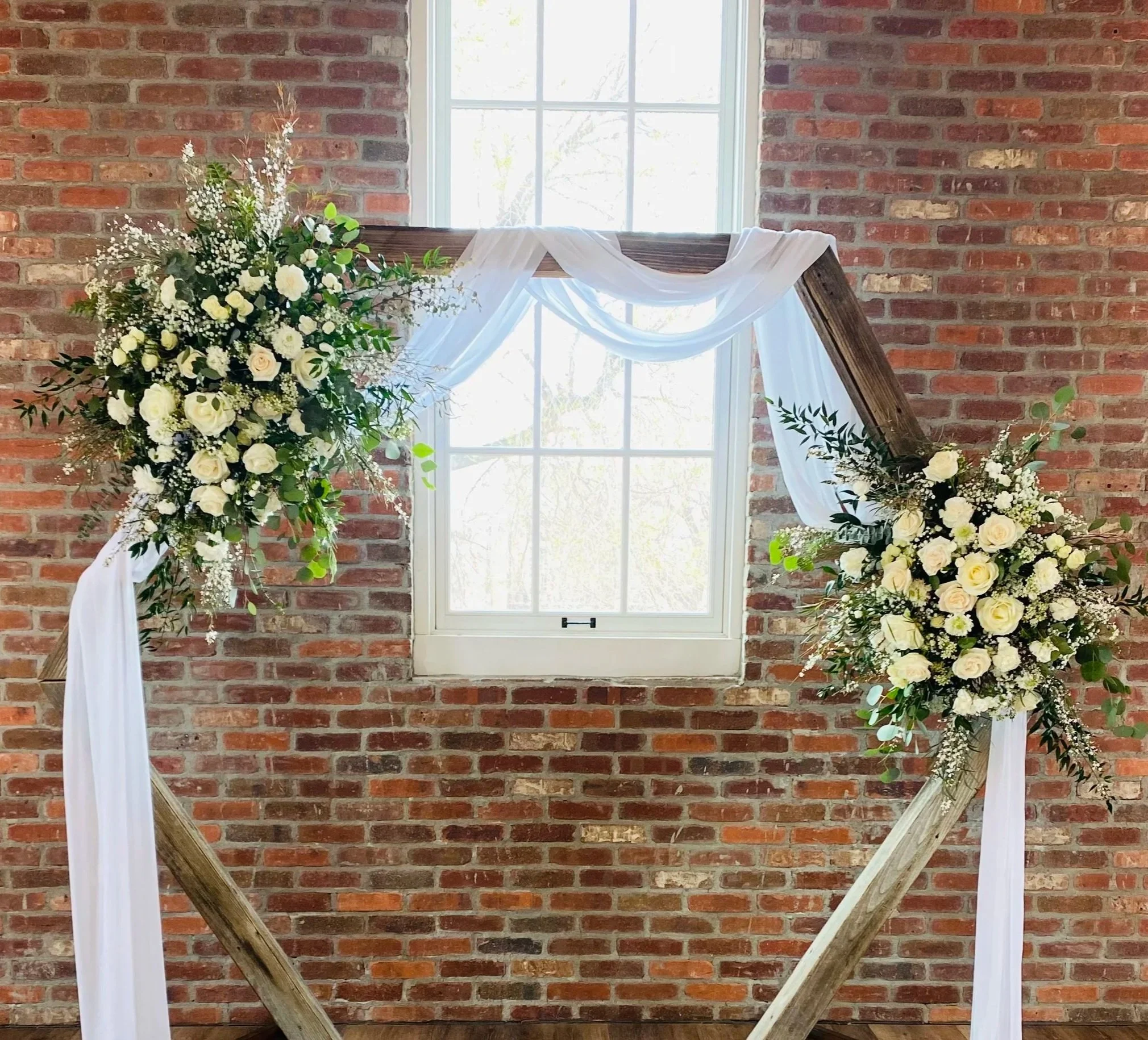 A wedding arch decorated with white fabric and white flowers, placed in front of a brick wall with a window letting in natural light.