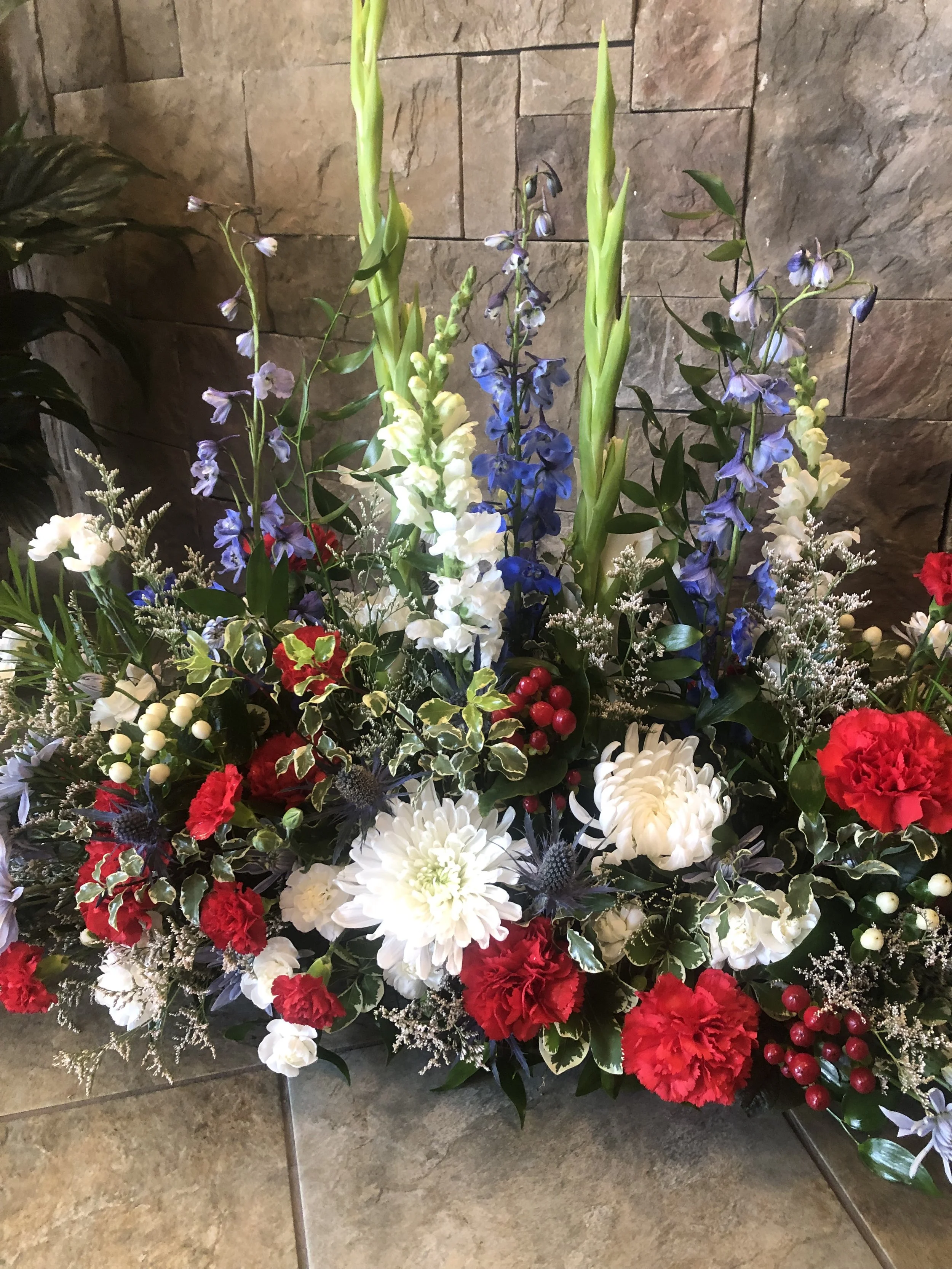 A sympathy flower arrangement with white, red, and blue flowers, greenery, and red berries, set against a textured stone wall on a tiled floor.