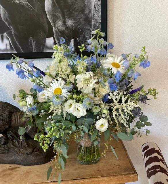 A large bridal bouquet of white, blue, and purple flowers with greenery in a glass vase on a wooden table, with a black and white horse photograph on the wall behind it.