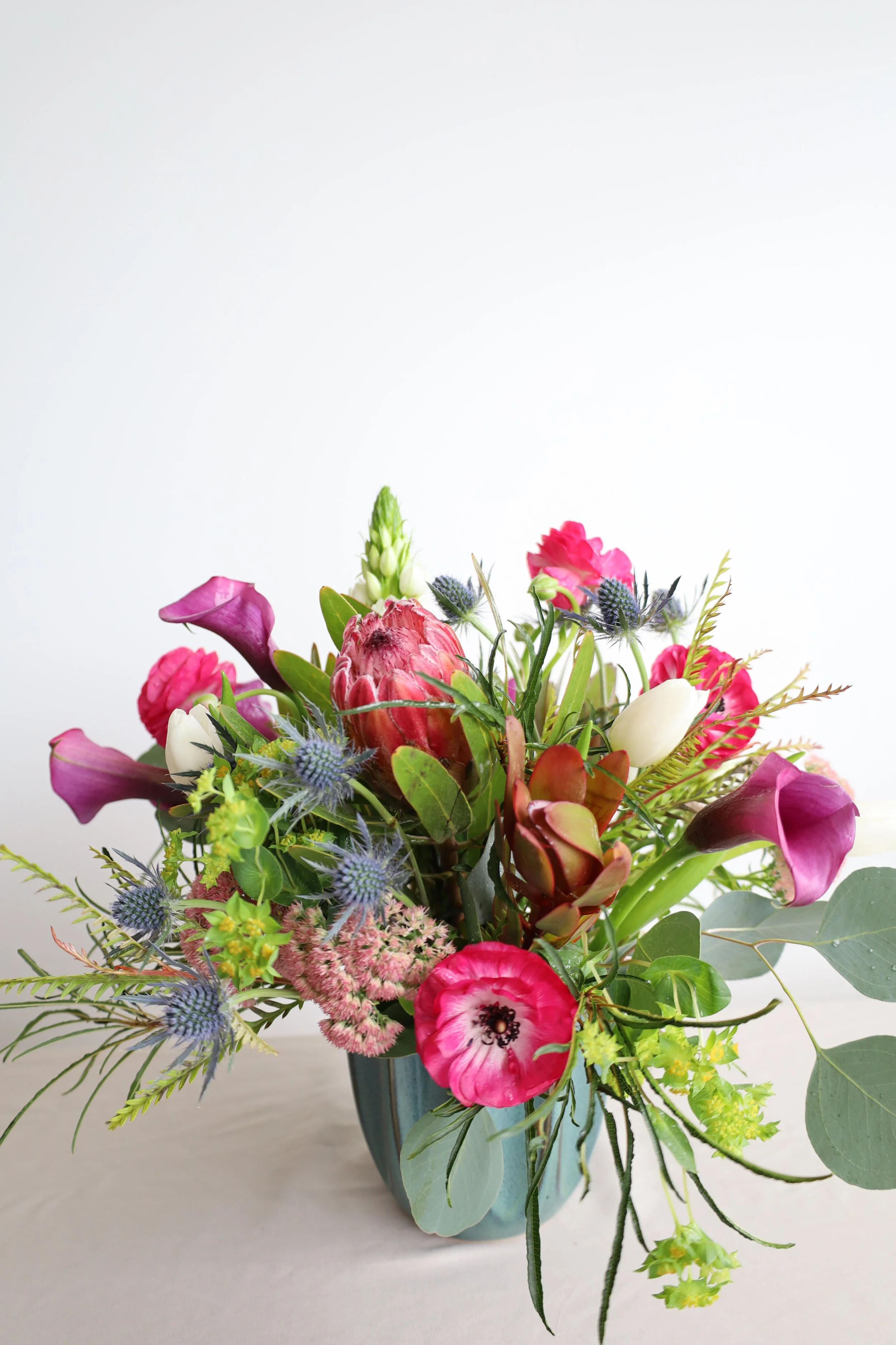 A bouquet of colorful flowers including pink, white, and purple blooms in a blue-green vase on a light surface against a white background.
