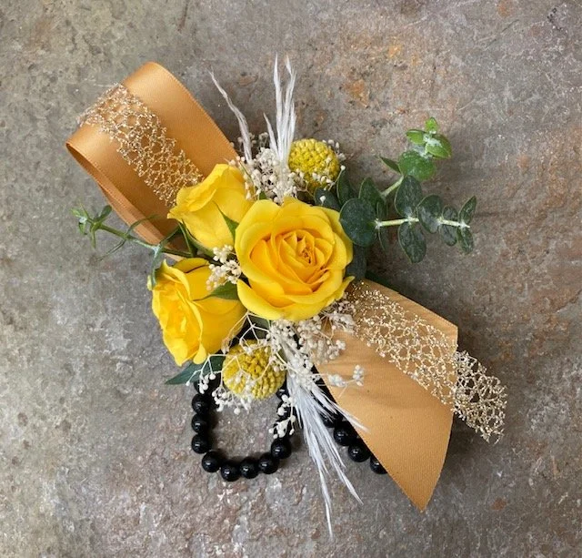 A wrist corsage with yellow roses, decorative gold lace ribbon, greenery, and dried accents on a tile surface.