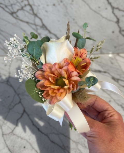 A pin-on corsage with a white rose, orange flowers, greenery, and white ribbon.
