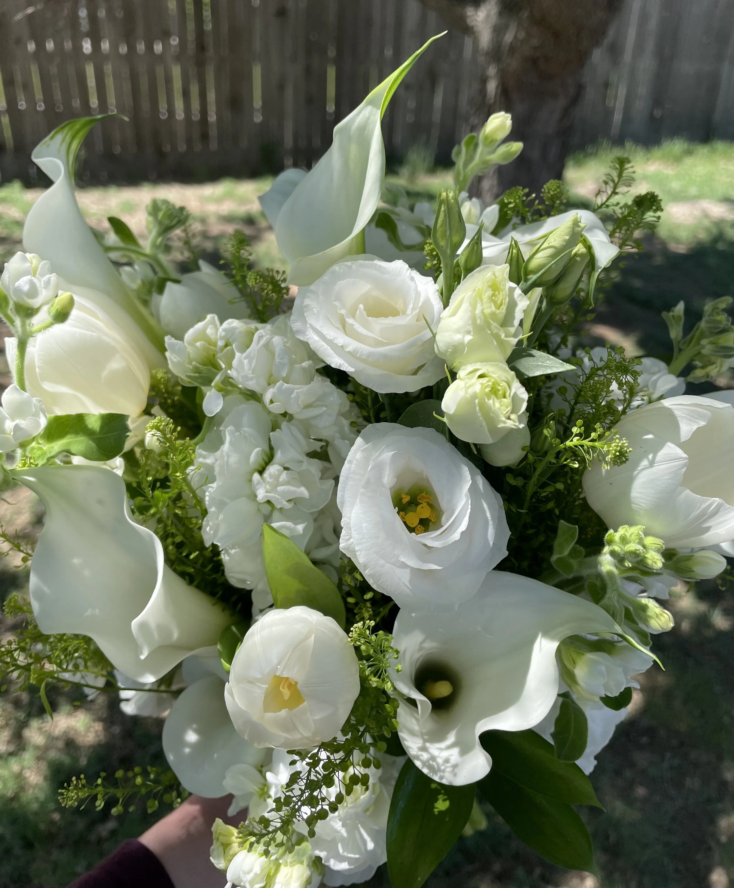 A bridal bouquet of white flowers, including lisianthus and calla lilies, held outdoors in daylight with a wooden fence in the background.