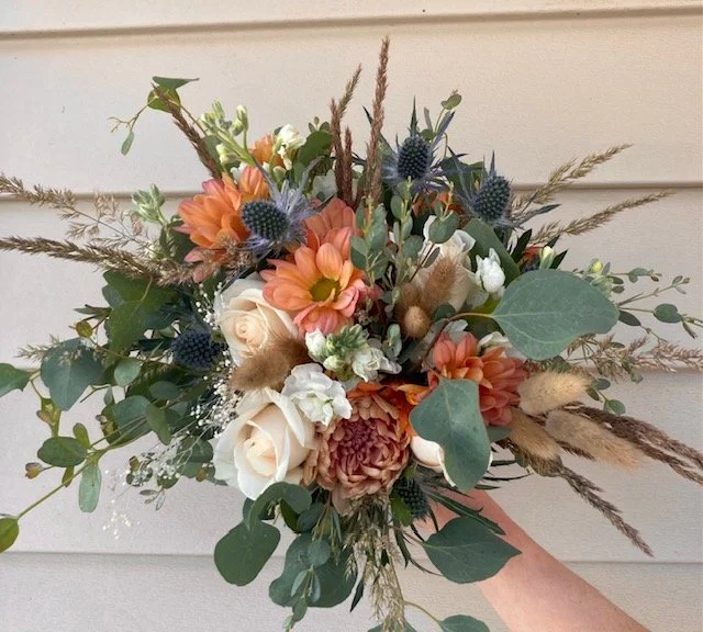 Hand-held bridal bouquet with white roses, peach daisies, blue thistles, and assorted greenery and dried grasses.