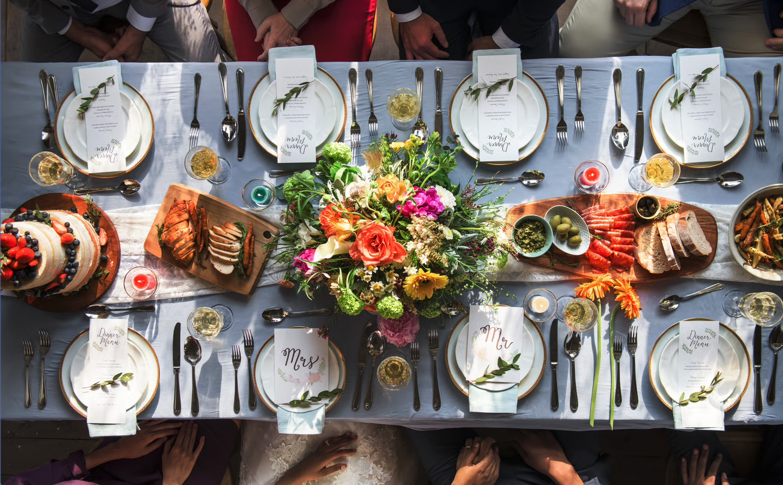 A beautifully set dining table with a centerpiece of colorful flowers, plates with menus, and an assortment of food including sliced bread, meats, and a cake topped with berries. Glasses of white wine and candles are also on the table, with guests seated around it.