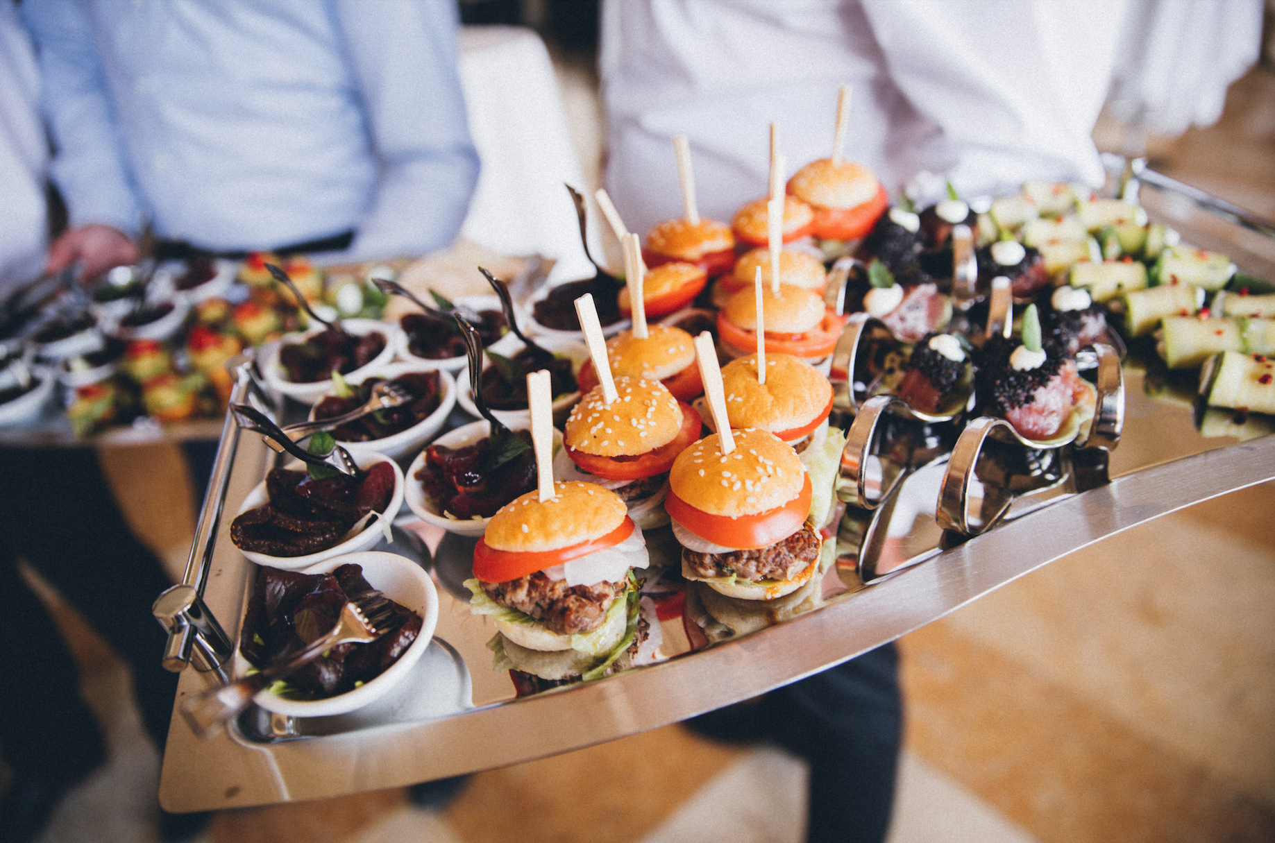 Tray of mini burgers with sesame seed buns, lettuce, tomato, and meat patty, served with small cups of salads or fruit, at a catered event.