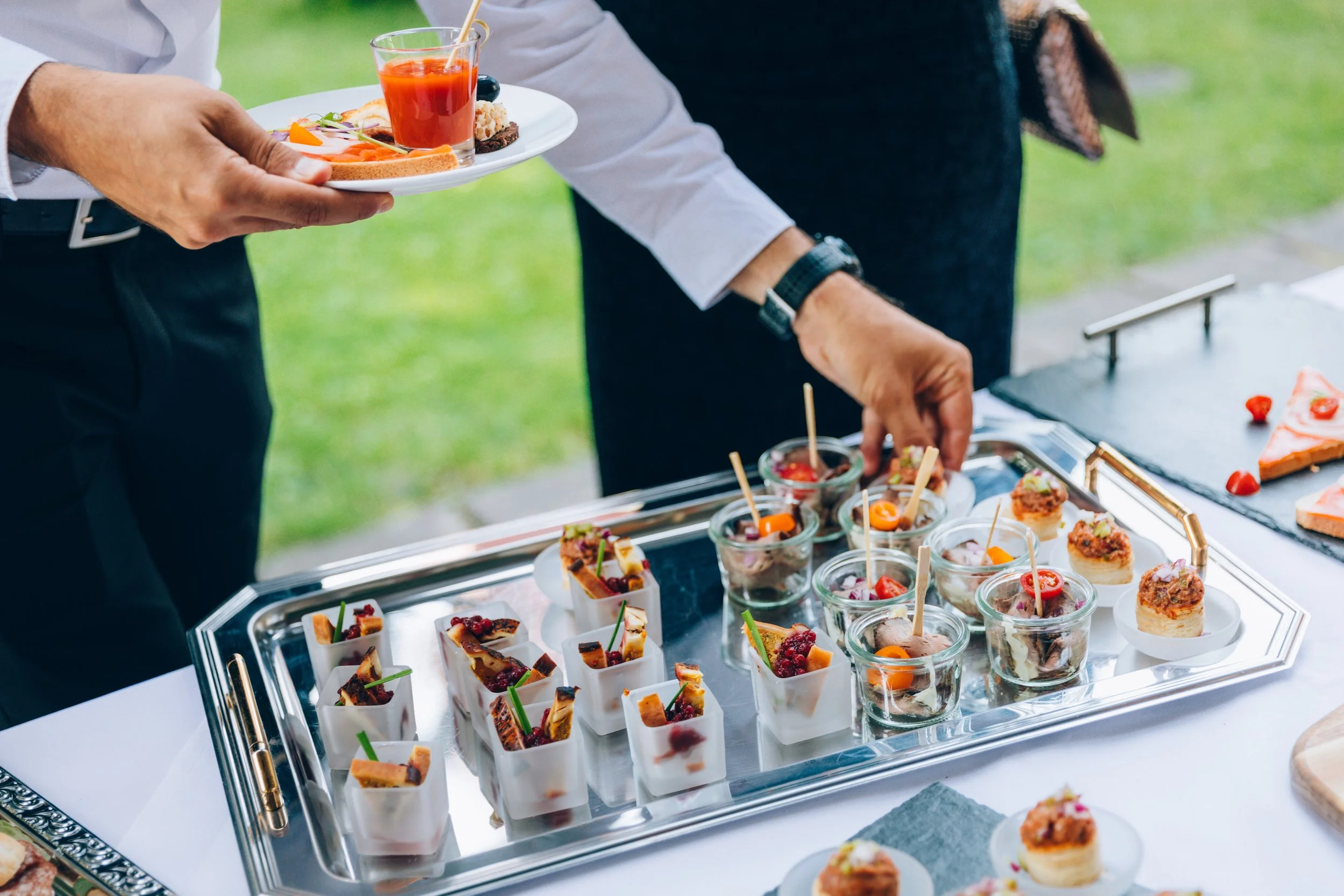 Person holding a white plate with small appetizers and a glass of red cocktail, at an outdoor buffet with various bite-sized foods on silver trays.