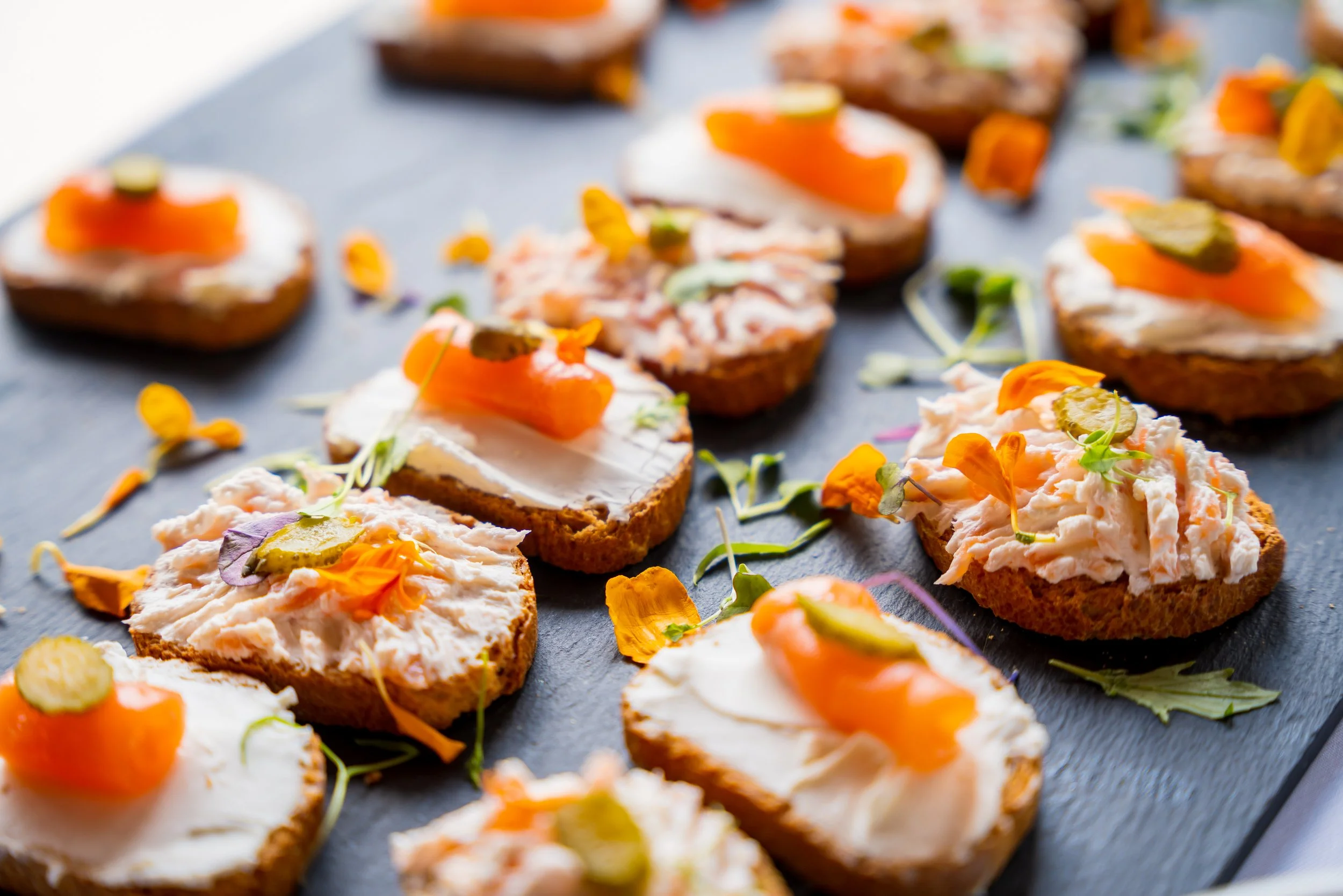 Assorted finger foods on a black slate serving platter, including toasted bread with creamy spread, smoked salmon, pickles, and edible flower garnishes.