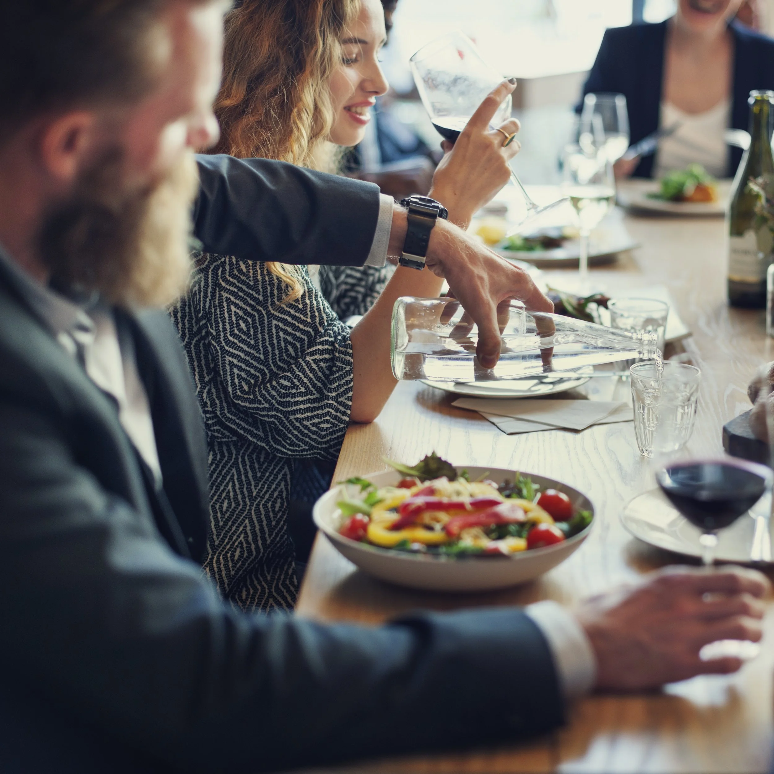 People sitting at a dining table during a social gathering, with food and drinks, including a woman holding a glass of red wine.
