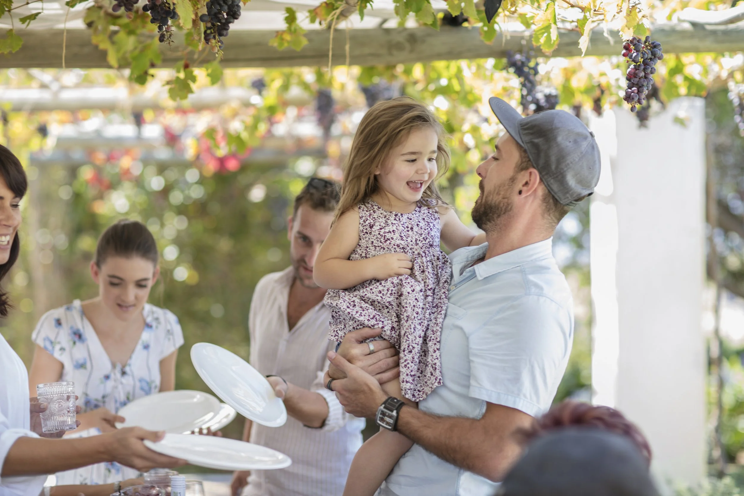 A man holding a young girl in a purple floral dress at a gathering outdoors under a canopy of grapevines. The girl is smiling and looking at the man, while other people are sitting at tables around them.
